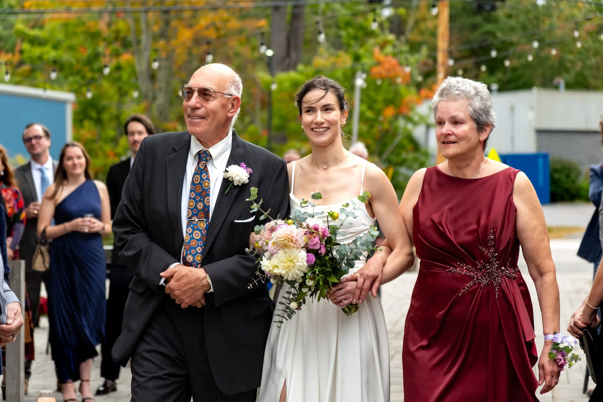 Victoria smiling as she walks down the aisle between her mom and dad at The Cellars at Allagash Brewing Company in Portland Maine fall wedding photography VMG Productions