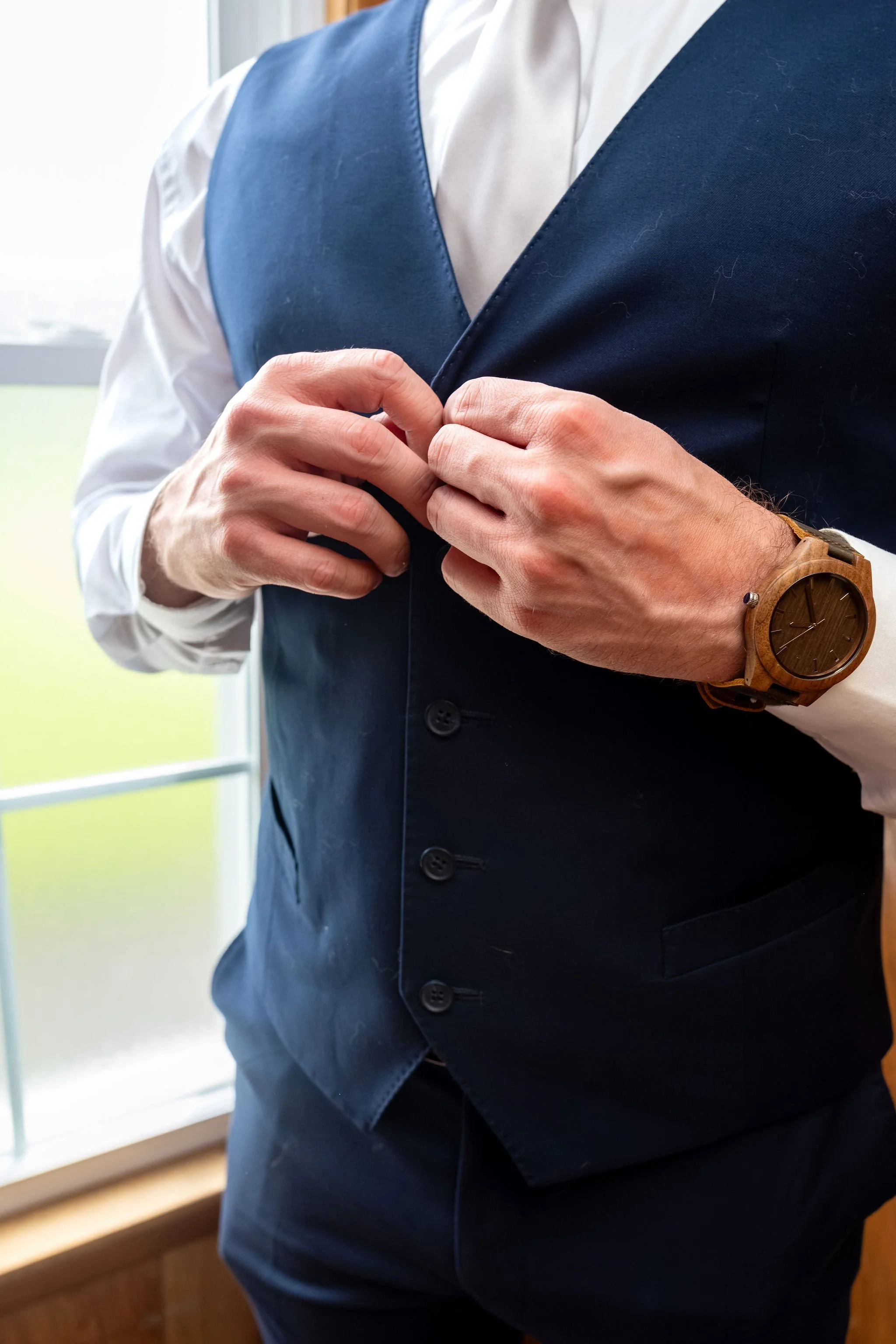 Beth and Cameron groom buttoning his navy vest while getting ready at Augusta Country Club Manchester Maine spring wedding photography VMG Productions