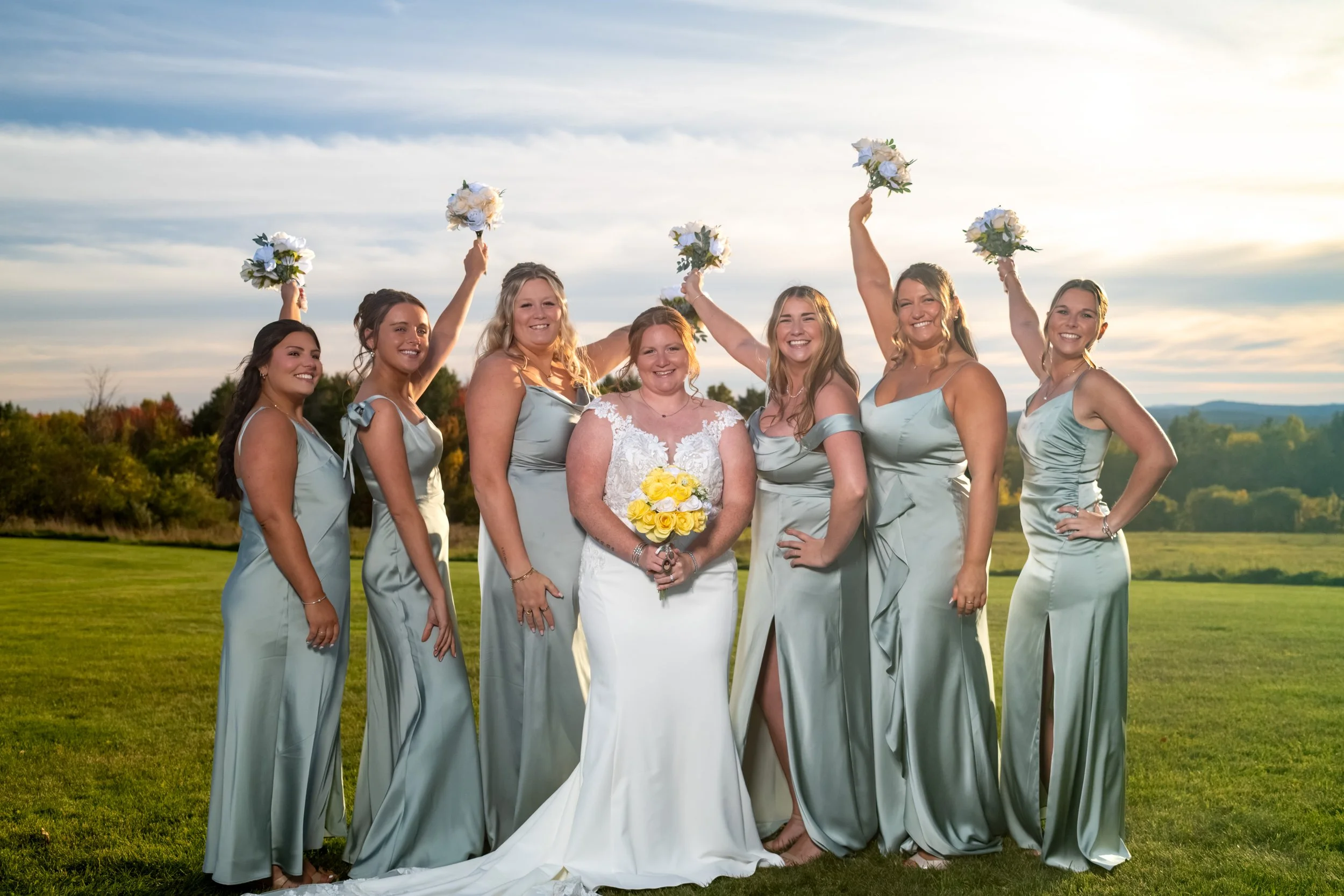 Erika with her bridesmaids in sage dresses cheering with their bouquets raised high on the lawn at the Maine Wedding Barn and Event Center in Minot Maine fall wedding photography VMG Productions