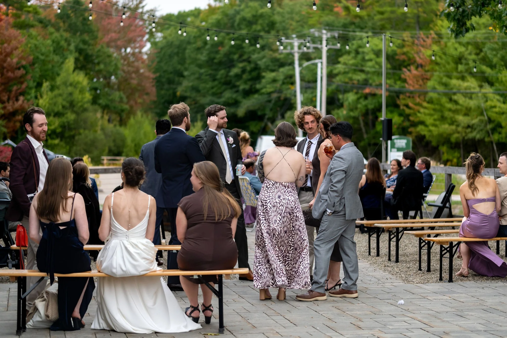 Victoria sitting on a wooden bench among guests on the patio as the afternoon gets going at The Cellars at Allagash Brewing Company in Portland Maine fall wedding photography VMG Productions