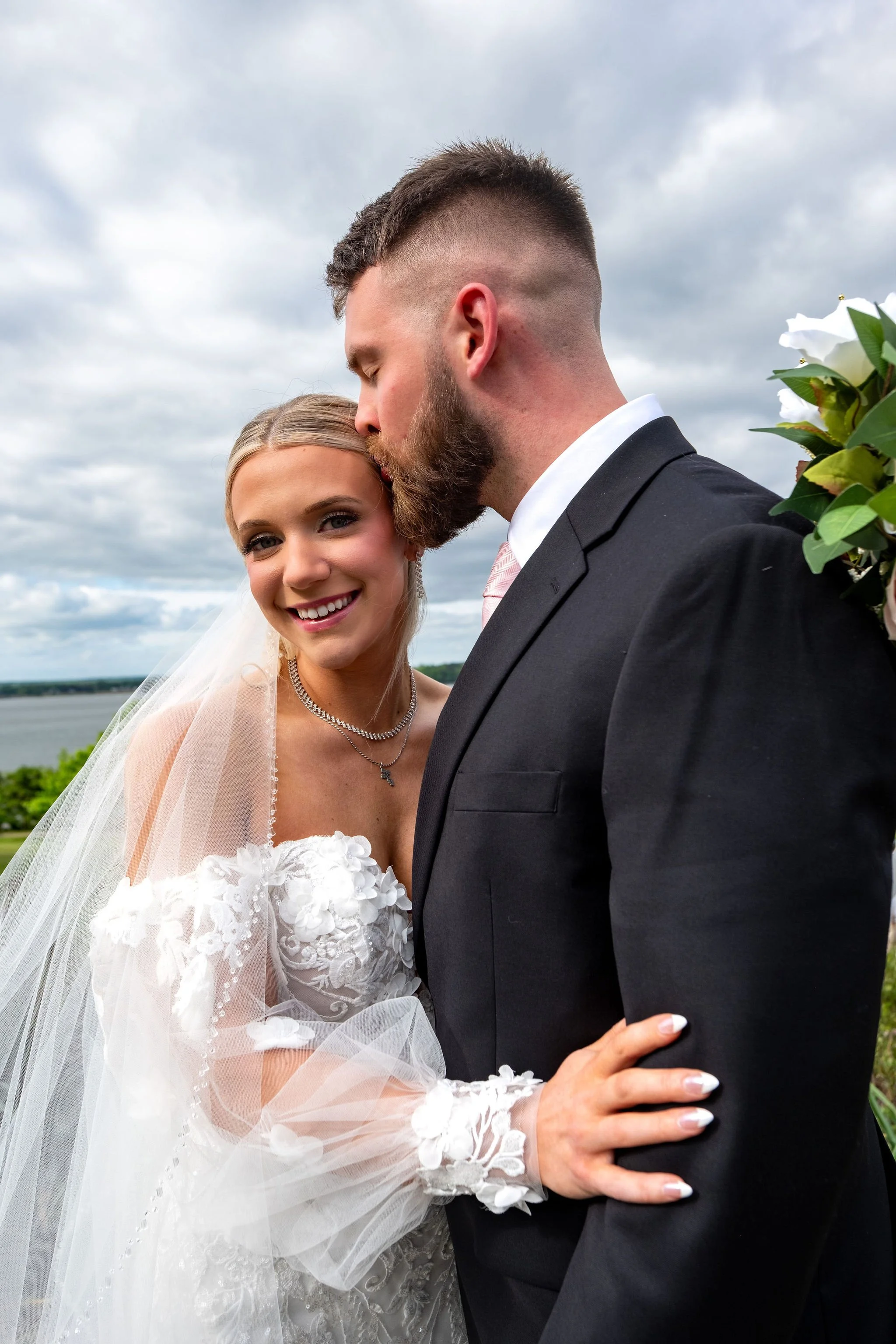 Jeremiah and Vienna bride smiles at camera while groom kisses her forehead during a quiet portrait moment at Eastern Promenade Portland Maine spring wedding photography VMG Productions
