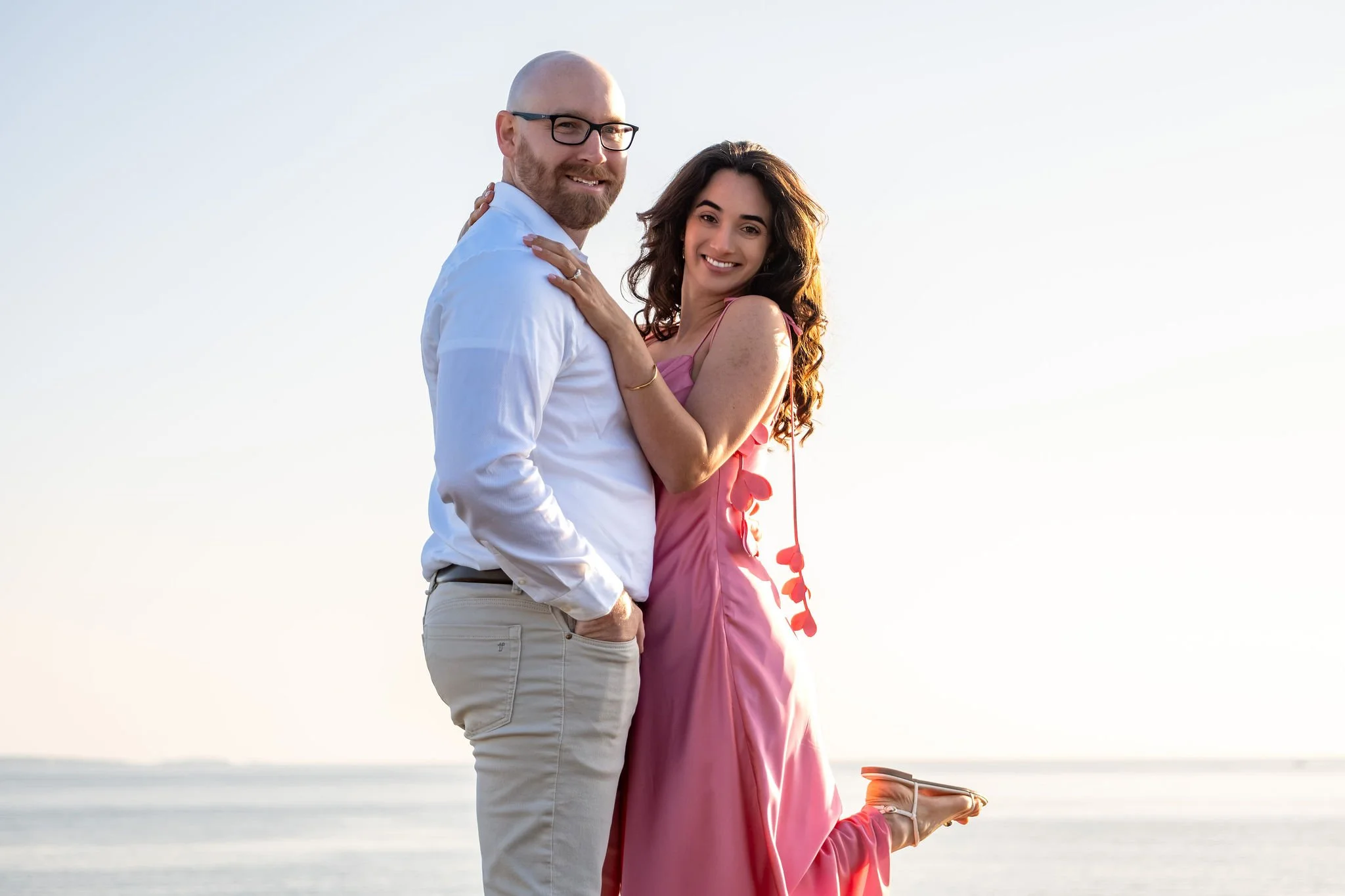 Zack and Helena smiling at the camera with Helena's foot kicked up behind her and the ocean behind them on Marginal Way in Ogunquit Maine sunrise proposal and engagement photography VMG Productions