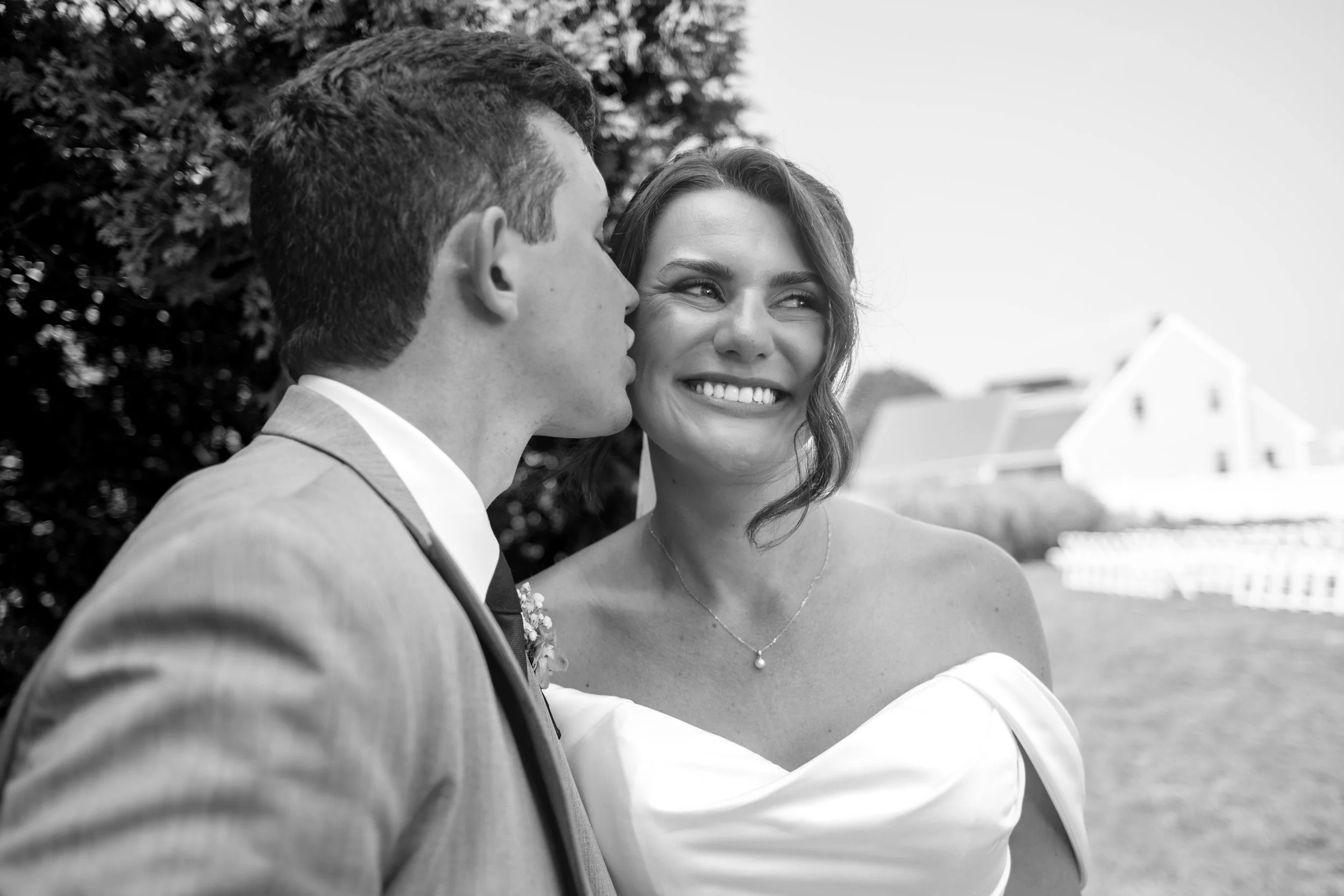 Anthony Pirolli kissing Caroline on the cheek as she smiles with the Union Bluff behind them in black and white in York Maine wedding photography VMG Productions