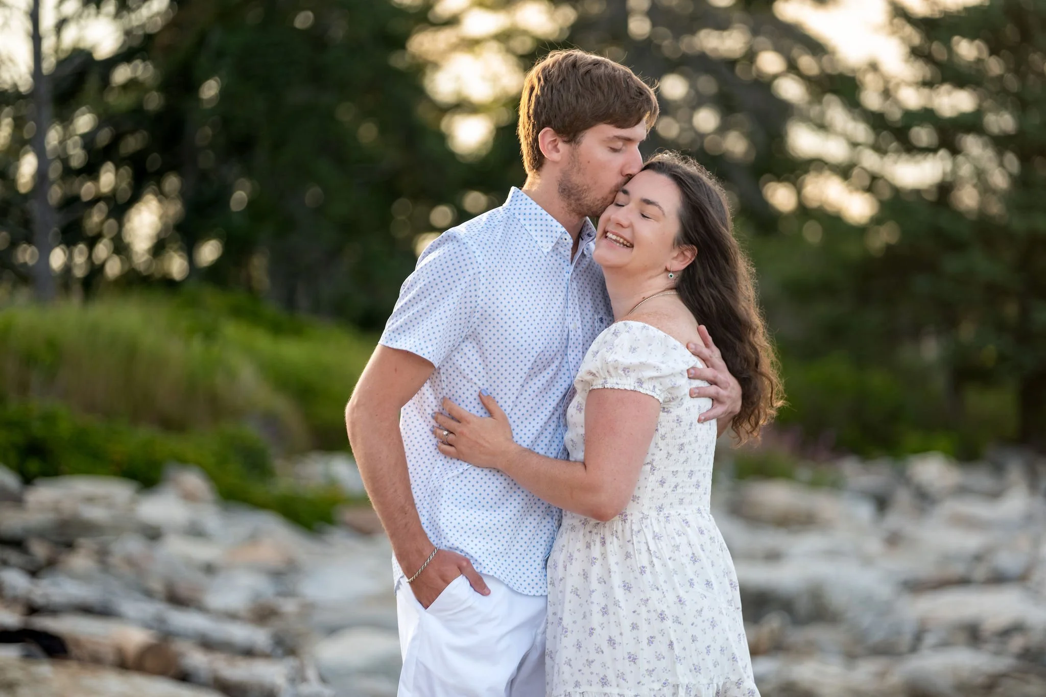 Lucas kissing Kirstie on the cheek on the ledges with the sun coming through the spruce at Reid State Park in Georgetown Maine engagement photography VMG Productions