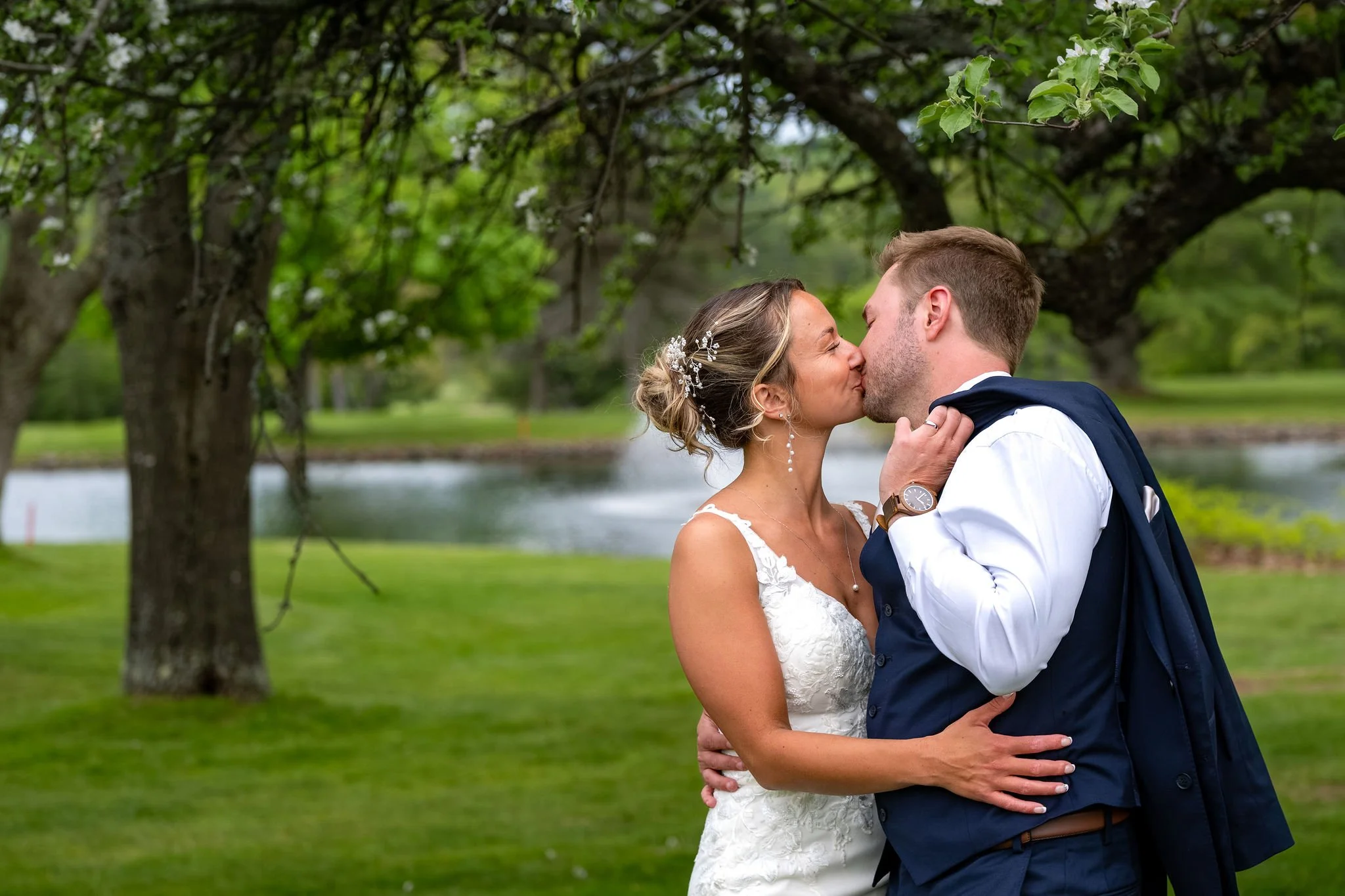 Beth and Cameron kissing beneath a flowering tree by the pond at Augusta Country Club Manchester Maine spring wedding photography VMG Productions