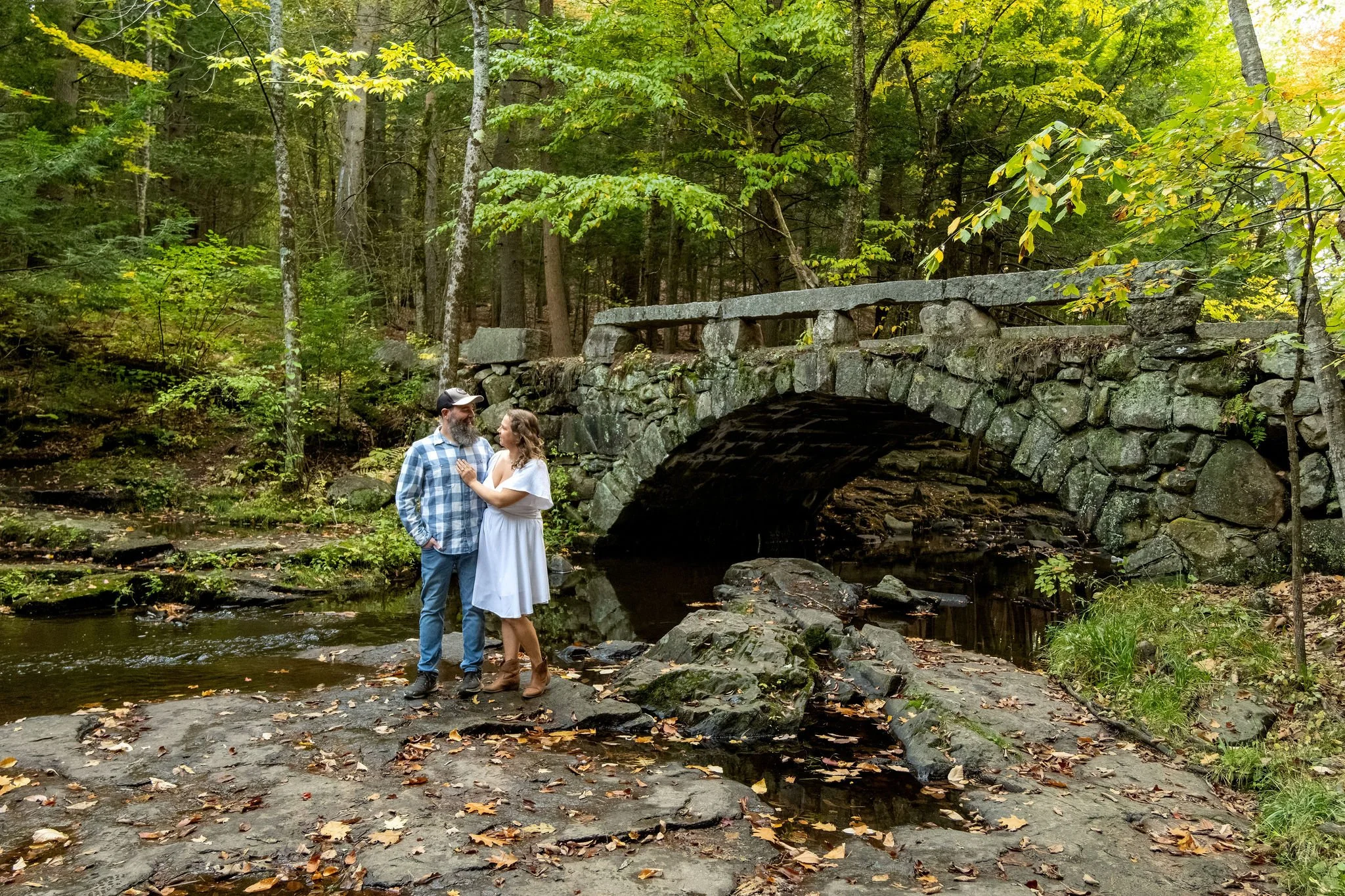 Wide view of Alison and Dave standing on the rocks beside the full stone arch bridge with the woods just starting to turn at Vaughan Woods in Hallowell Maine fall engagement photography VMG Productions