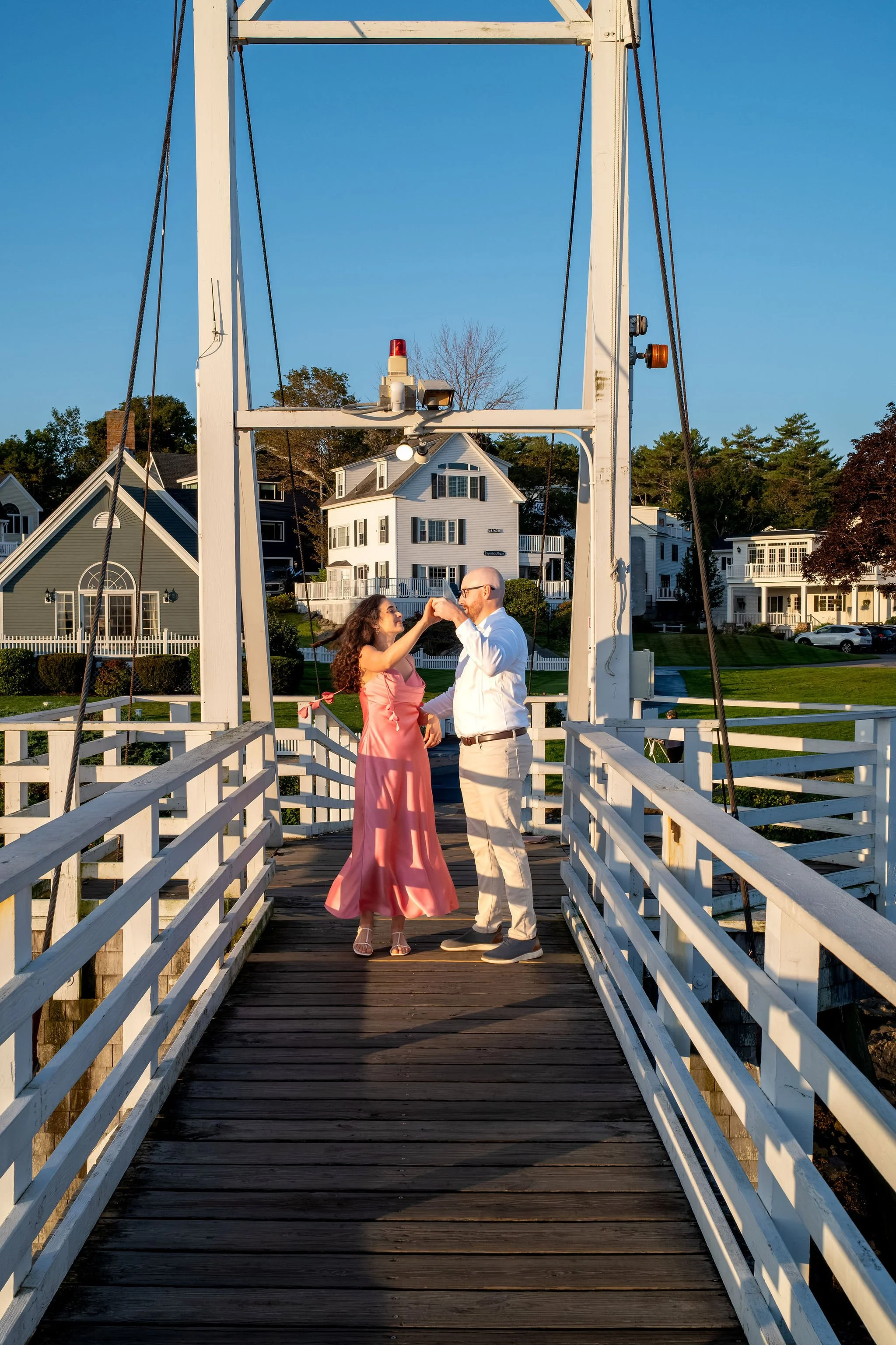 Zack and Helena dancing together in the middle of the white footbridge with the harbor houses behind them on Marginal Way in Ogunquit Maine sunrise proposal and engagement photography VMG Productions
