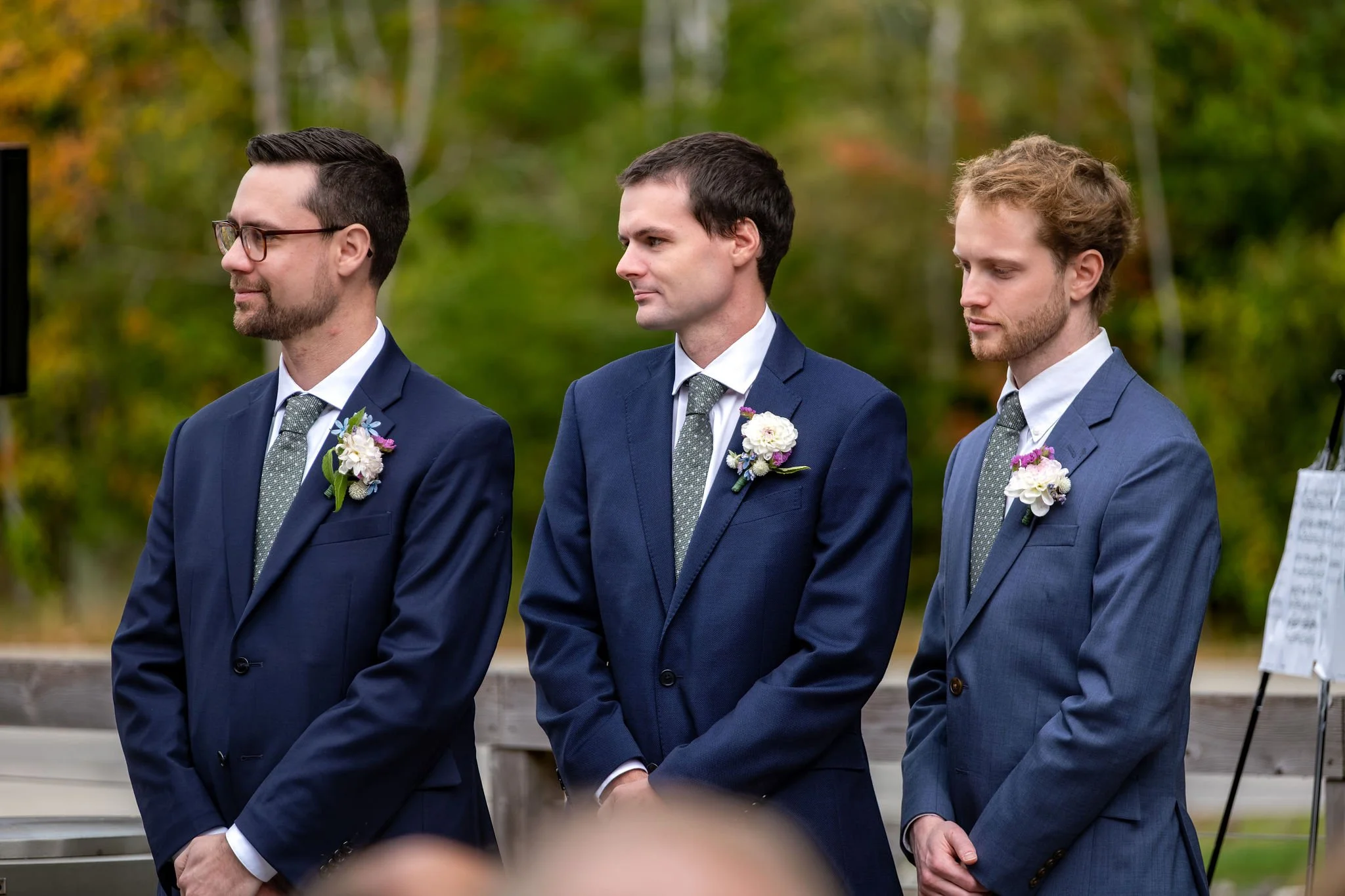 Three groomsmen in navy and blue suits watching the ceremony at The Cellars at Allagash Brewing Company in Portland Maine fall wedding photography VMG Productions