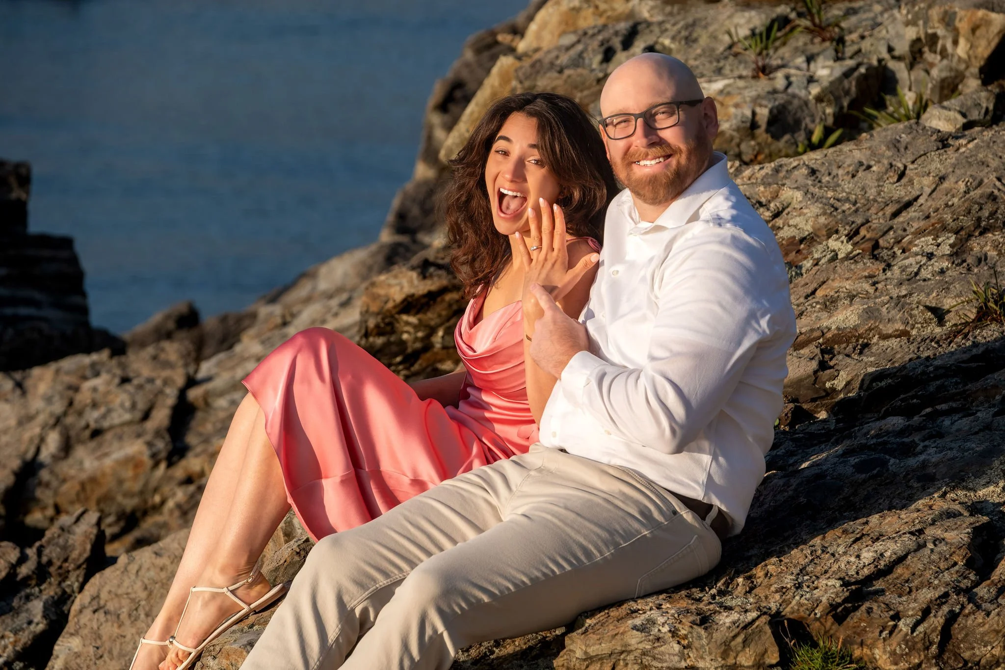 Helena holding up her ring with a huge excited smile as Zack grins beside her on the rocks on Marginal Way in Ogunquit Maine sunrise proposal and engagement photography VMG Productions