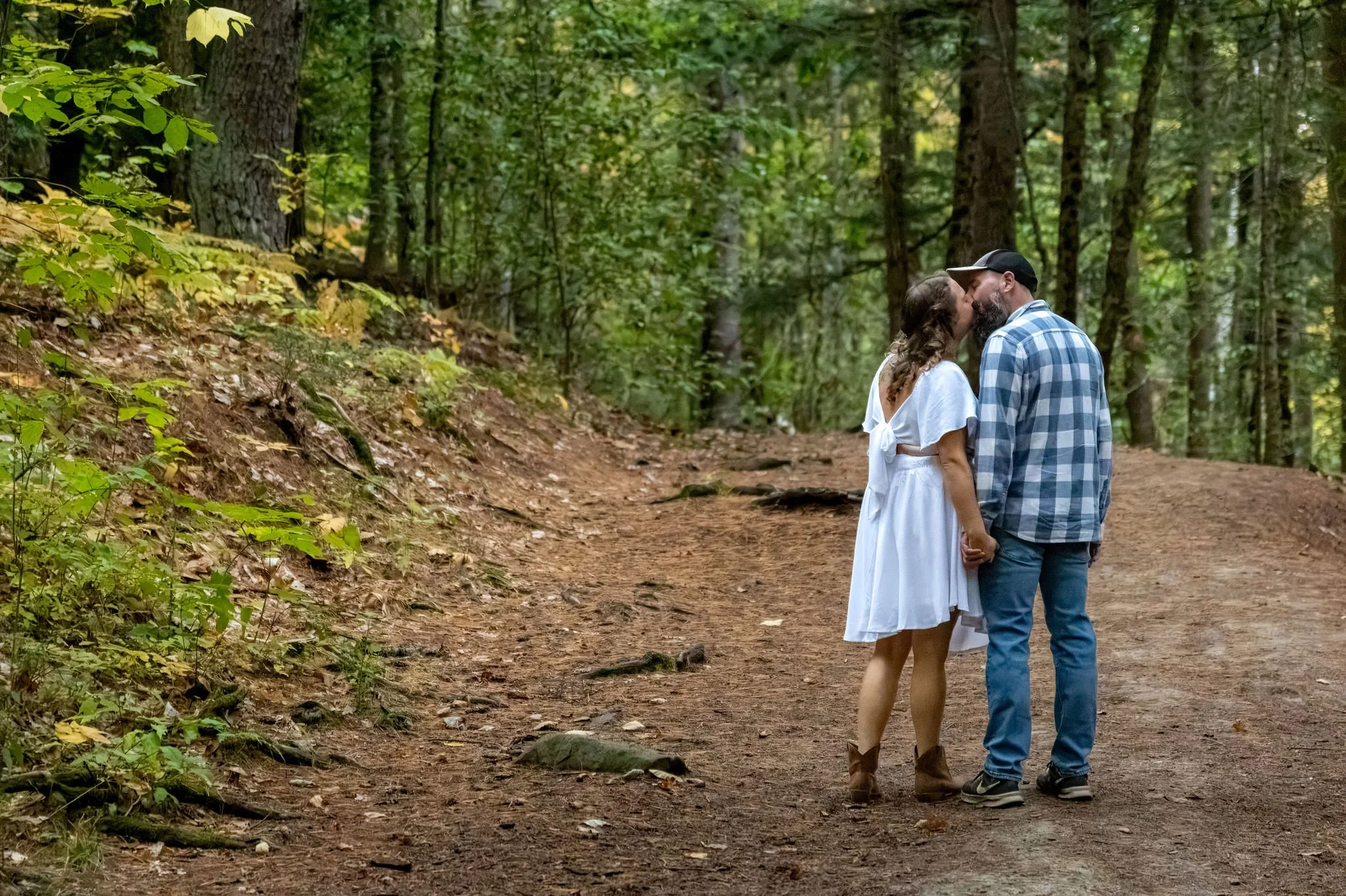 Alison and Dave kissing on the wooded trail with the ferns and leaves on the hillside behind them at Vaughan Woods in Hallowell Maine fall engagement photography VMG Productions