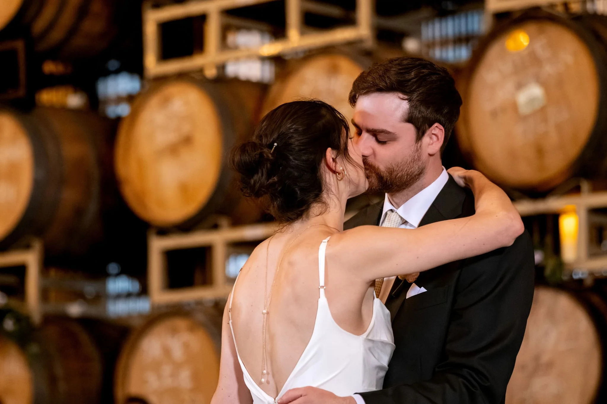 Victoria and Jack kissing during their first dance in front of the barrels at The Cellars at Allagash Brewing Company in Portland Maine fall wedding photography VMG Productions