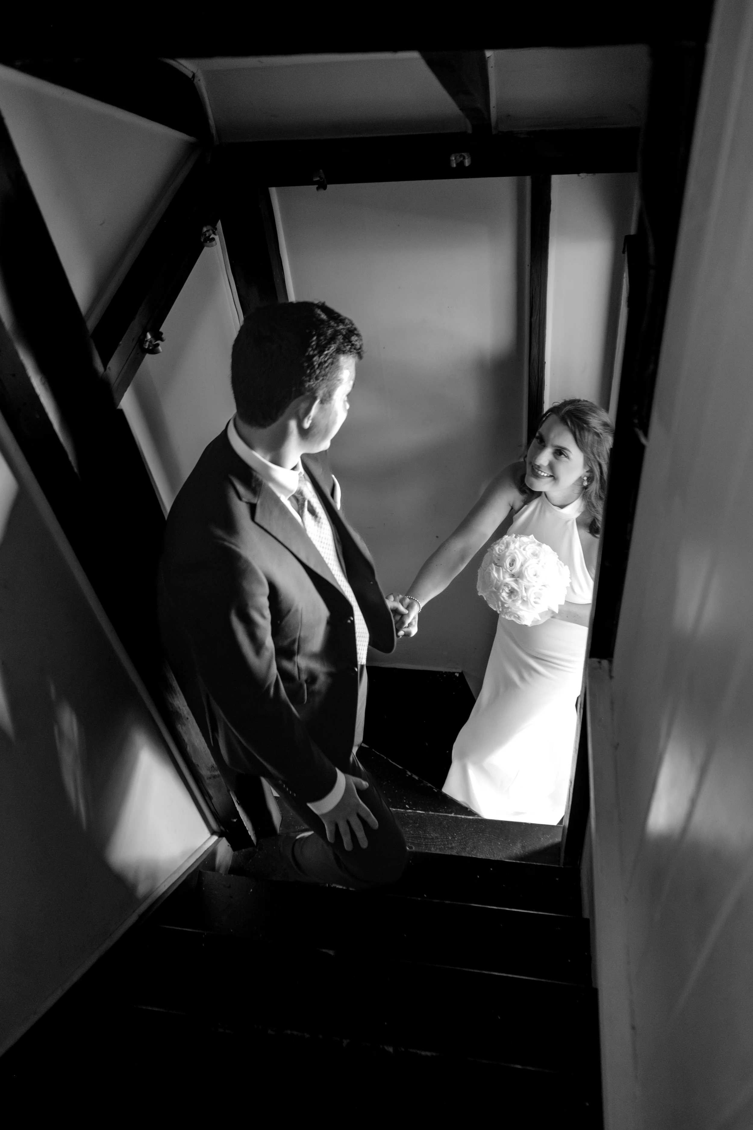 Caroline and Anthony Pirolli holding hands on the staircase looking back at each other in black and white at Star of the Sea in York Maine wedding photography VMG Productions