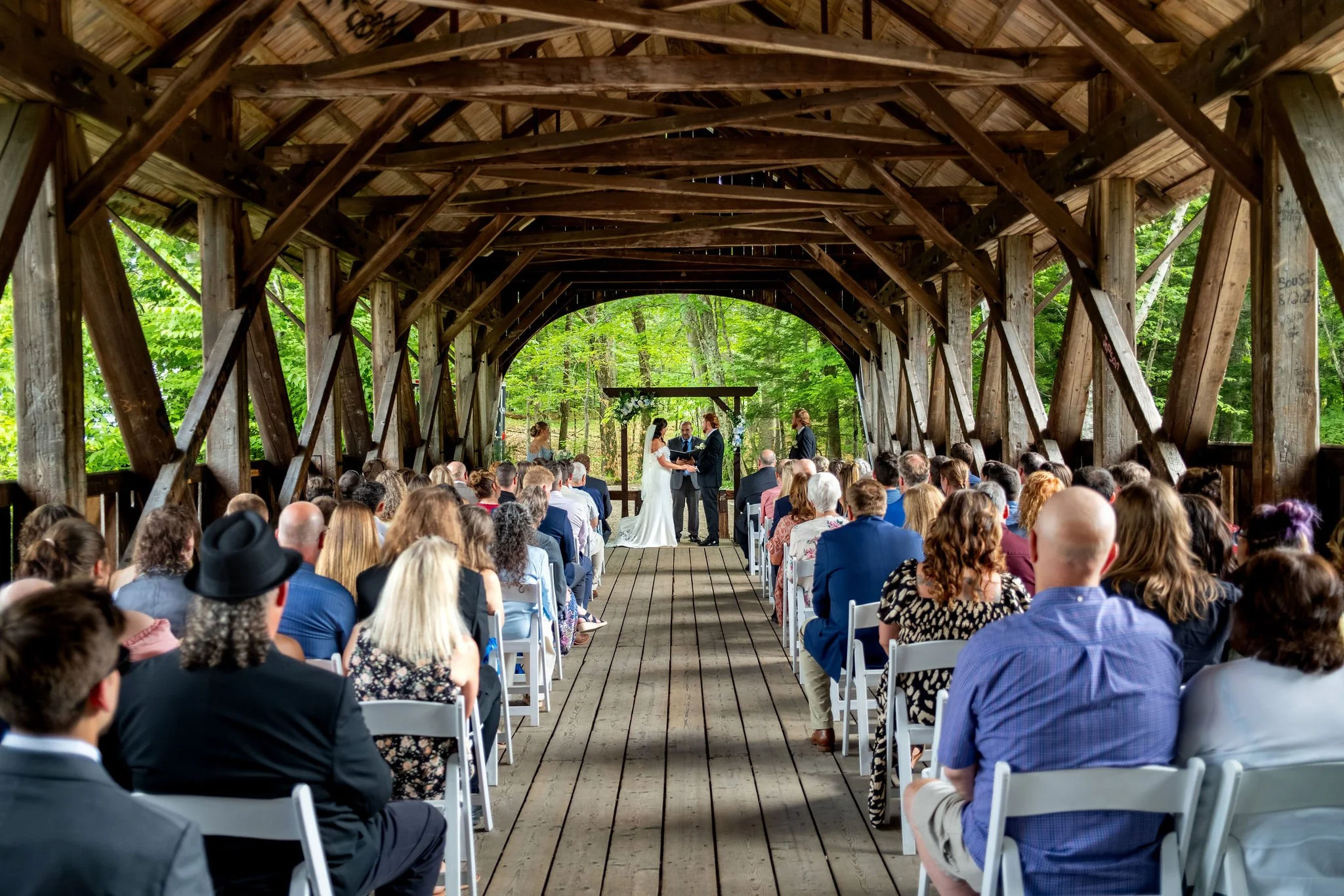 Alyssa and Colby wide view of the wedding ceremony inside the timber trusses at Sunday River Covered Bridge Newry Maine summer wedding photography VMG Productions