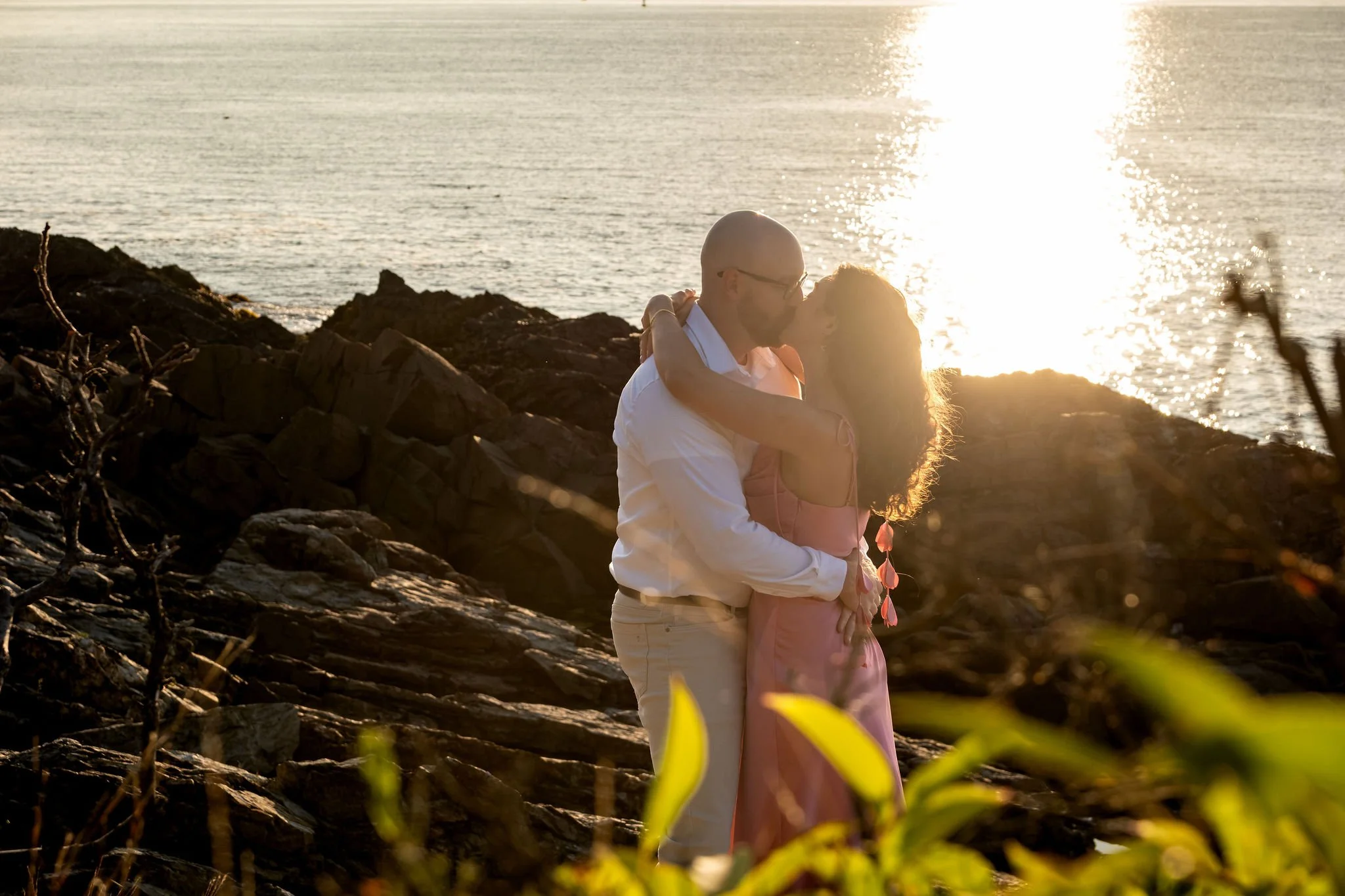 Zack and Helena embracing on the rocks with the sun sparkling across the ocean behind them on Marginal Way in Ogunquit Maine sunrise proposal and engagement photography VMG Productions