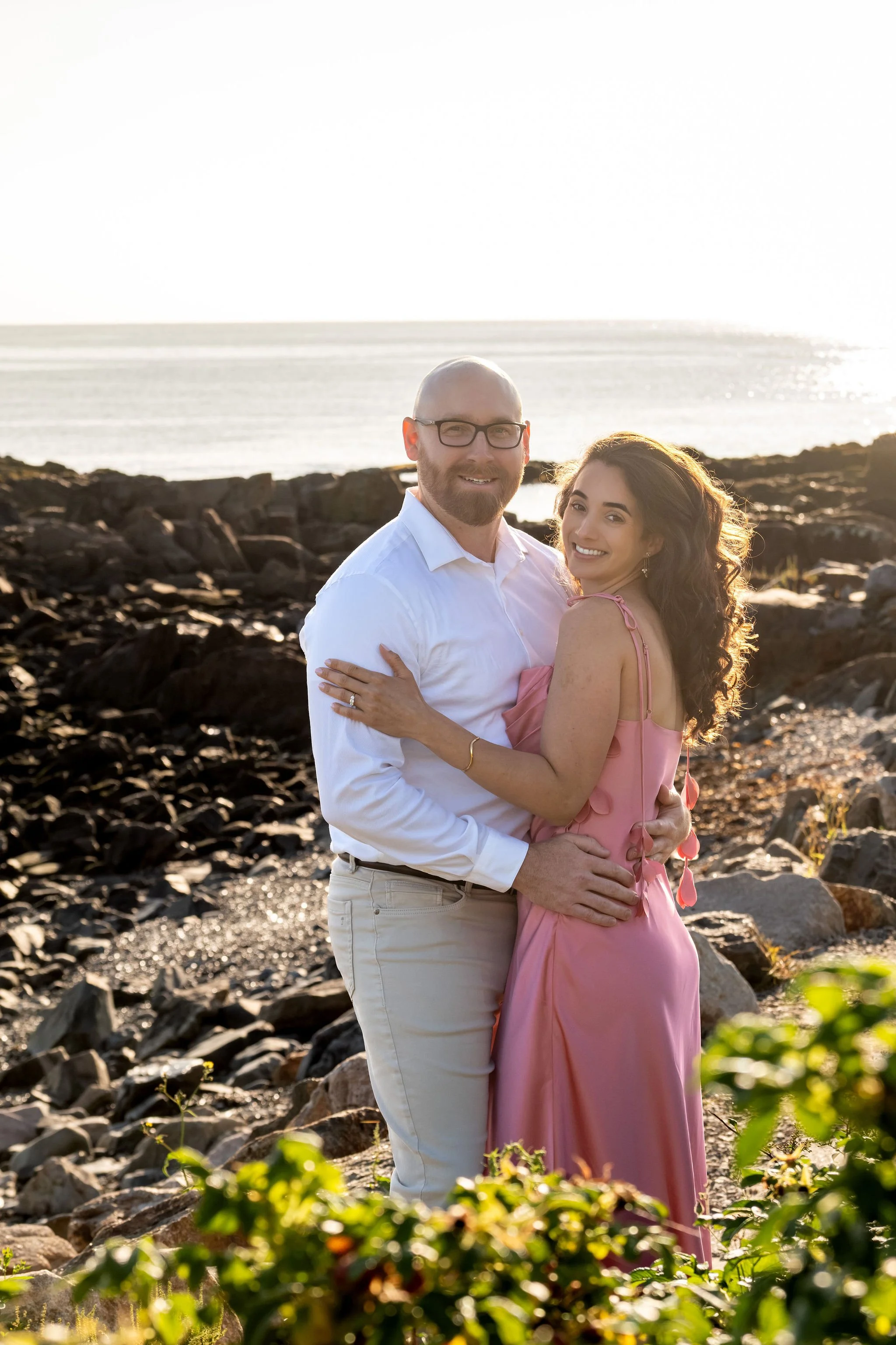 Zack and Helena smiling at the camera with their arms around each other and the dark rocky shore behind them on Marginal Way in Ogunquit Maine sunrise proposal and engagement photography VMG Productions