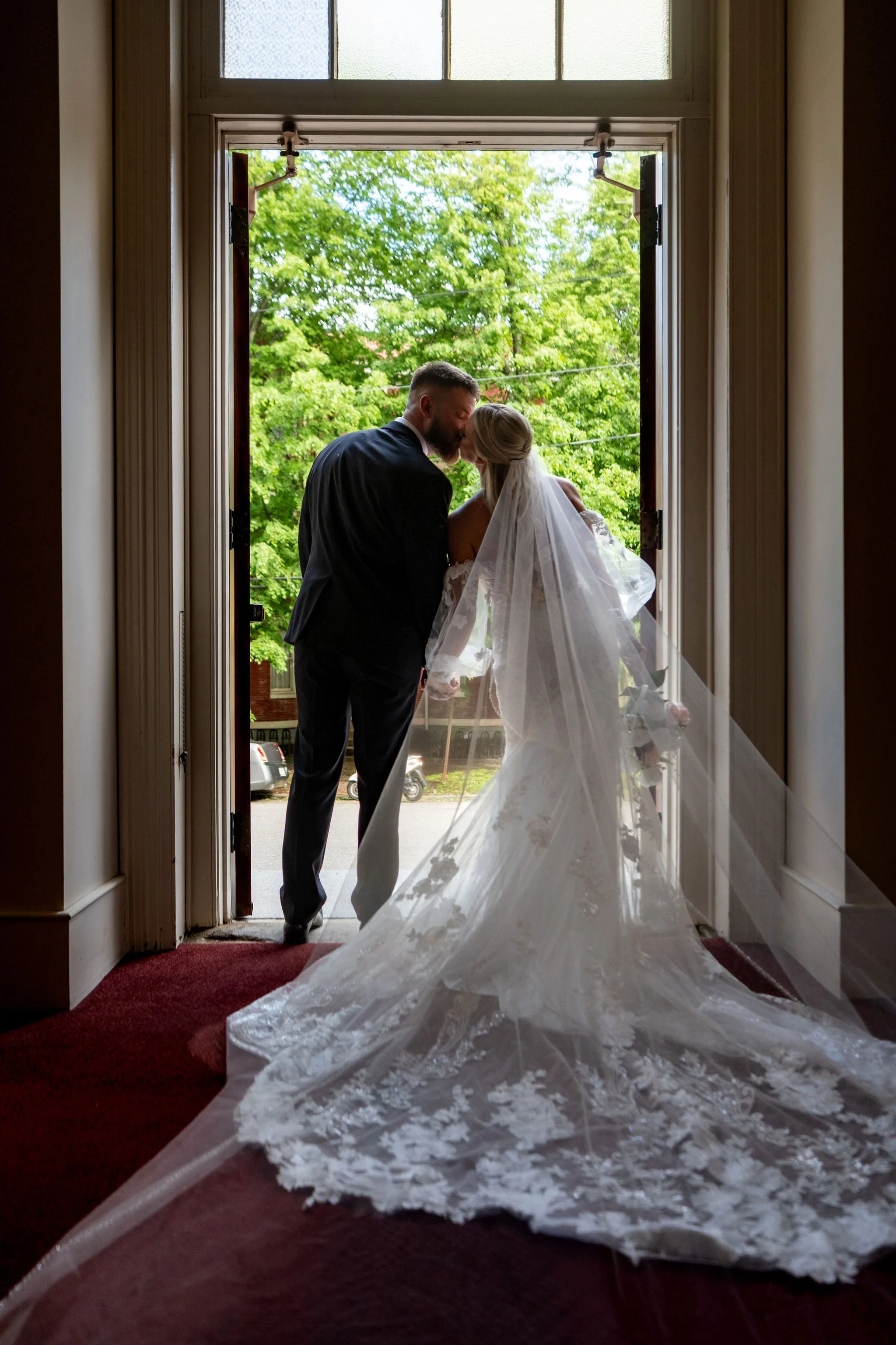 Jeremiah and Vienna couple shares a kiss framed by the open doorway while the veil cascades across the floor at Holy Trinity Greek Orthodox Church Portland Maine spring wedding photography VMG Productions