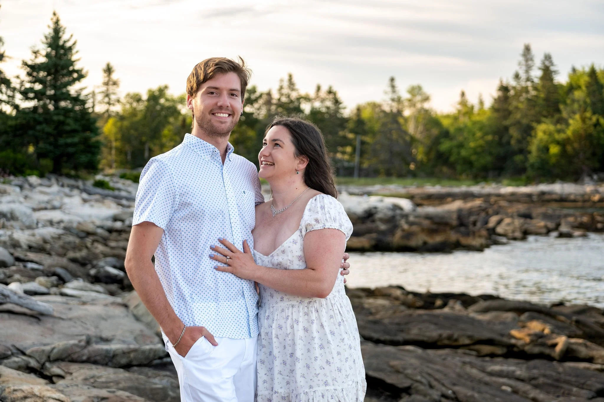 Kirstie and Lucas standing together on the granite ledges with the cove and spruce trees behind them at Reid State Park in Georgetown Maine engagement photography VMG Productions