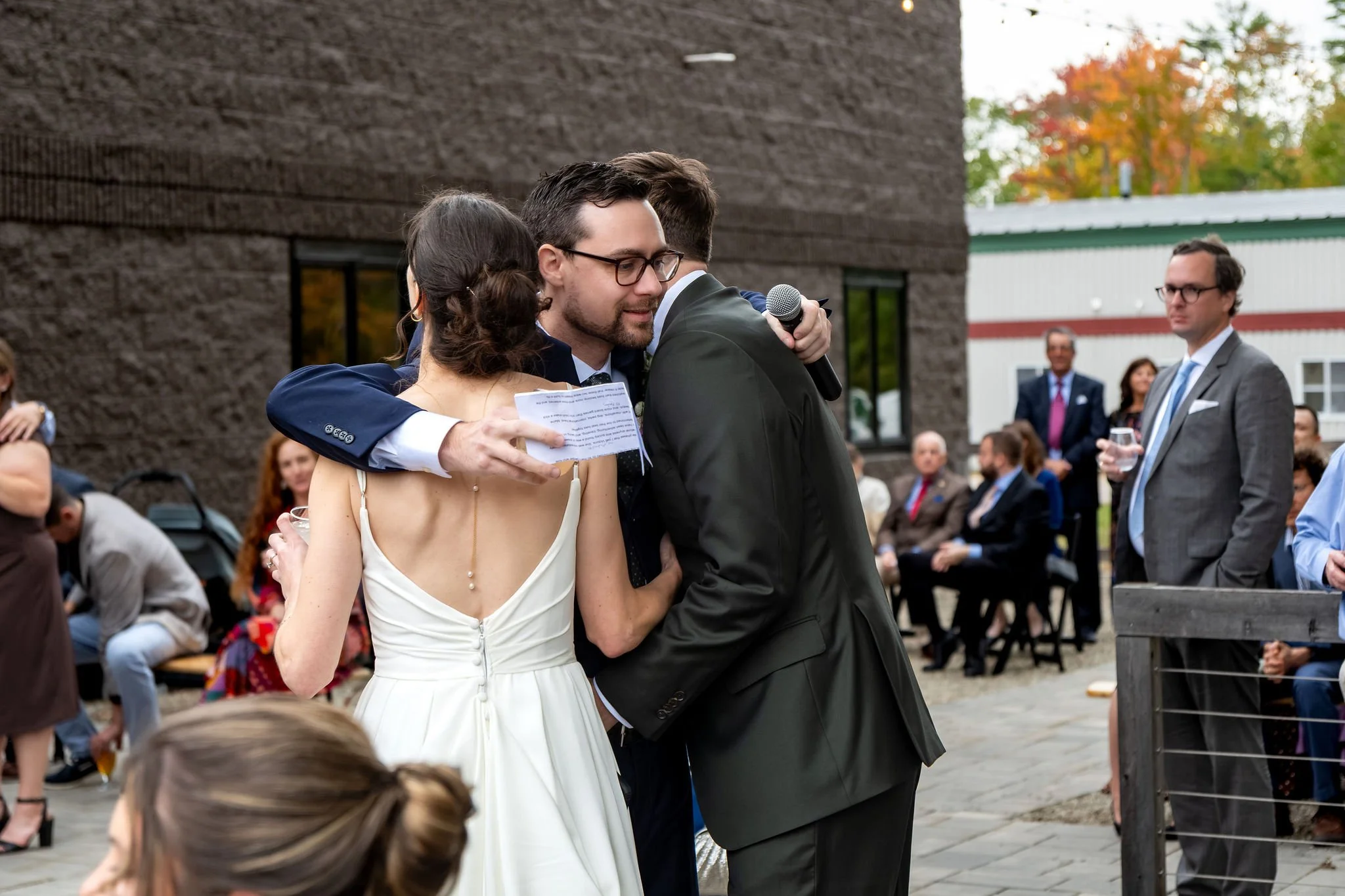 One of the groomsmen hugging Victoria and Jack after his toast with the microphone still in hand at The Cellars at Allagash Brewing Company in Portland Maine fall wedding photography VMG Productions