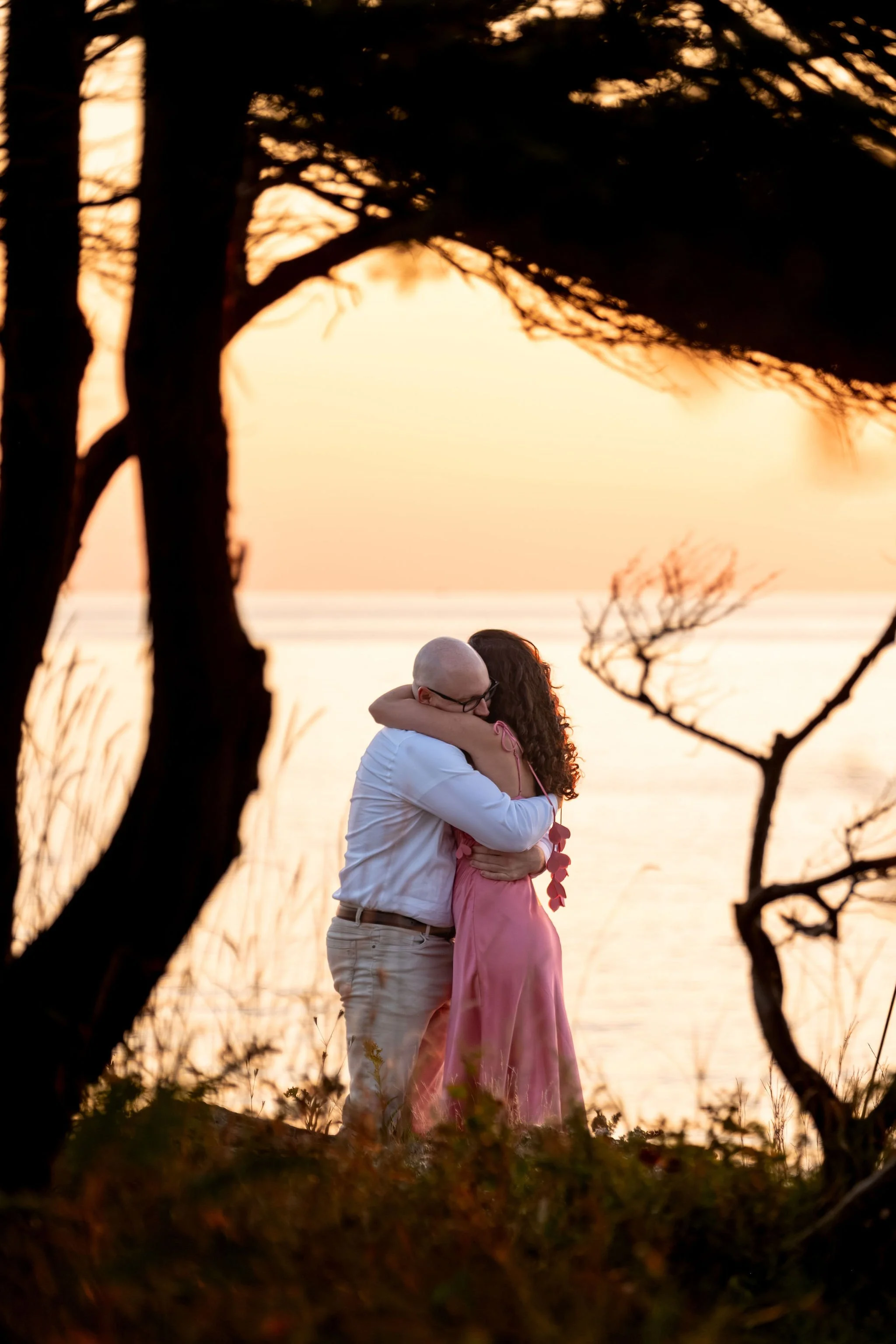 Zack and Helena hugging tightly beside the silhouetted tree on Marginal Way in Ogunquit Maine sunrise proposal and engagement photography VMG Productions
