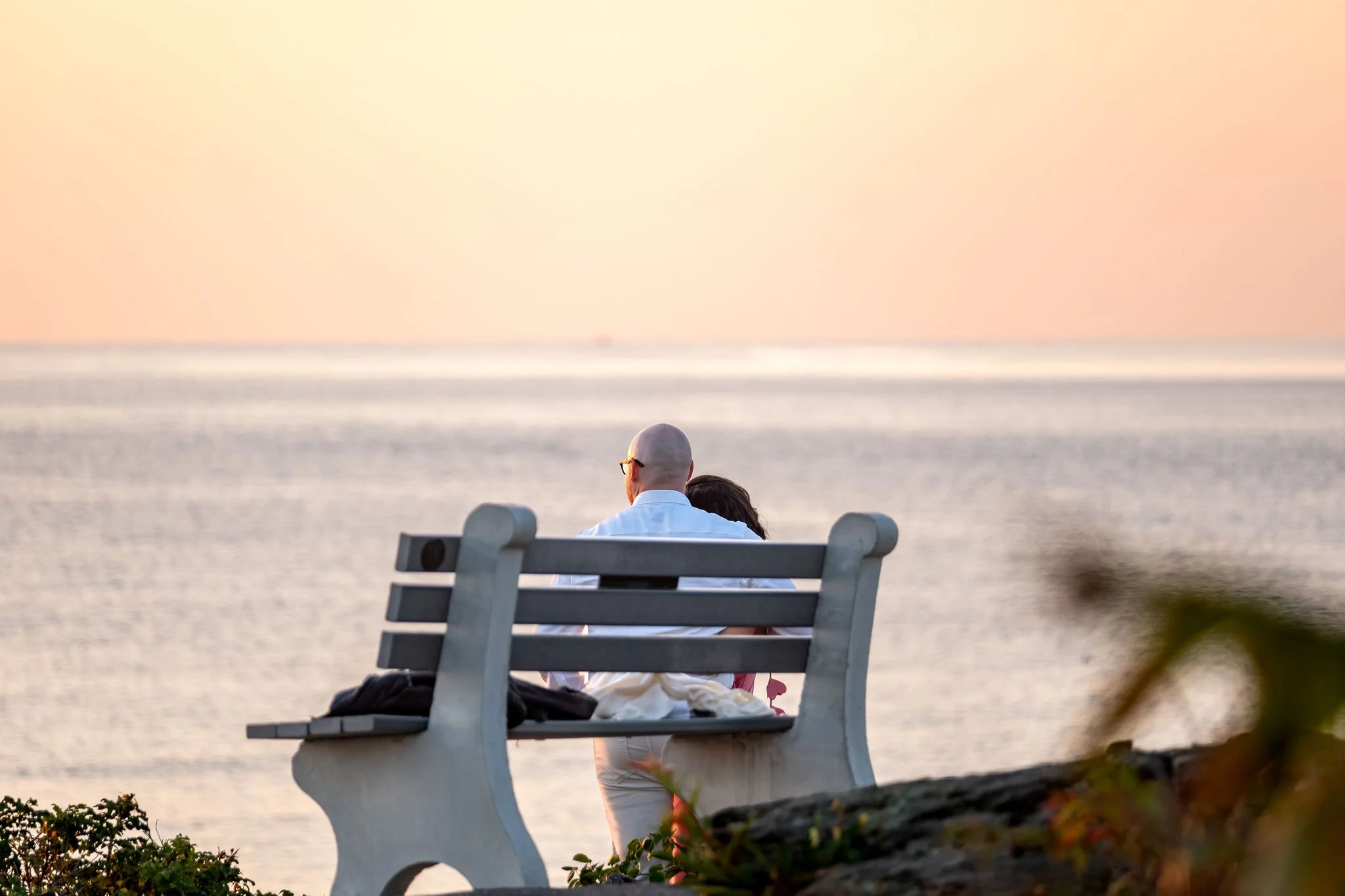 Zack and Helena sitting close on a white bench watching the calm ocean glow in the early light on Marginal Way in Ogunquit Maine sunrise proposal and engagement photography VMG Productions