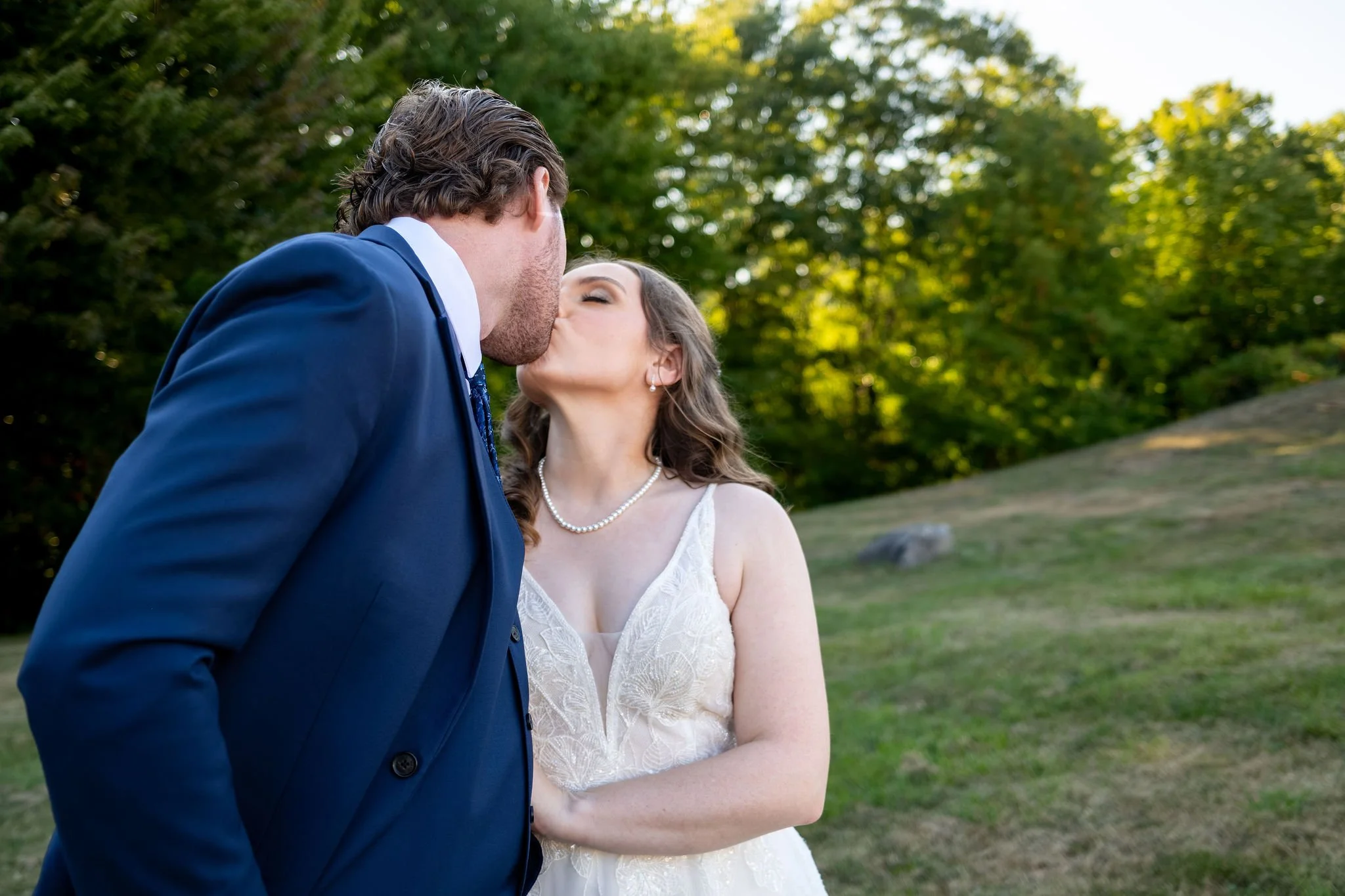 Zachary and Hollie kissing in the field with the tree line lit gold behind them at The Lucerne Inn in Dedham Maine summer wedding photography VMG Productions