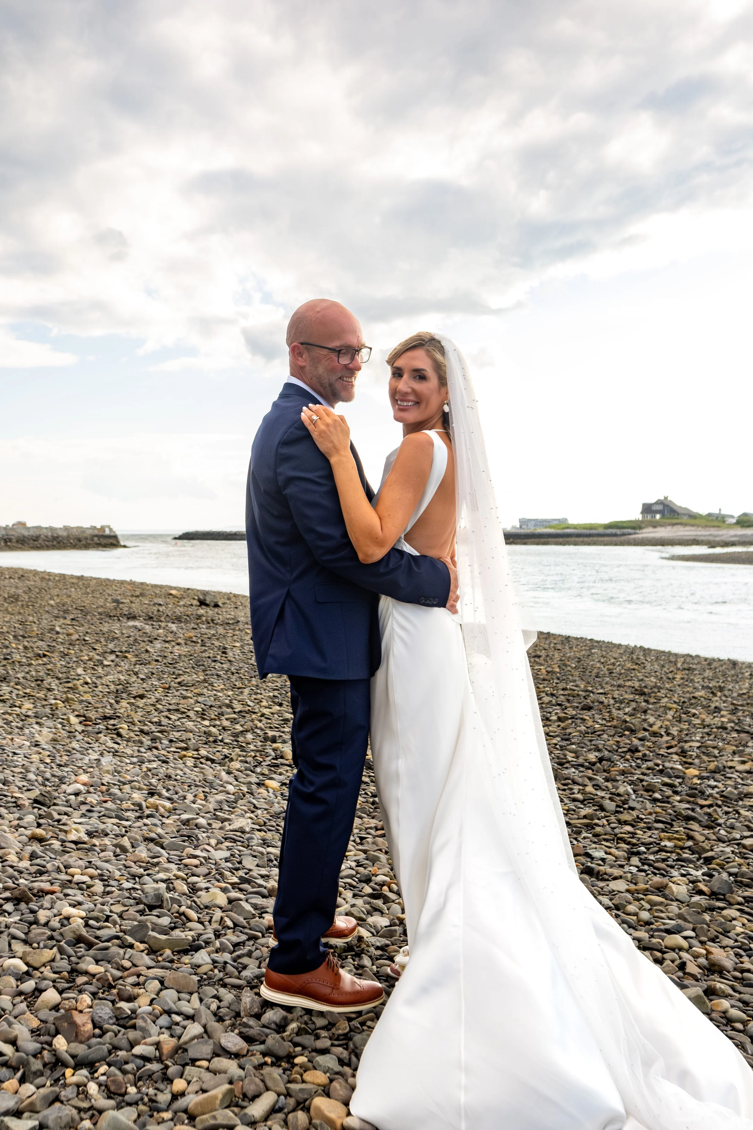 Ashley looking back over her shoulder as she and Marc embrace on the pebble beach after their ceremony at The Breakwater Inn in Kennebunkport Maine summer wedding photography VMG Productions