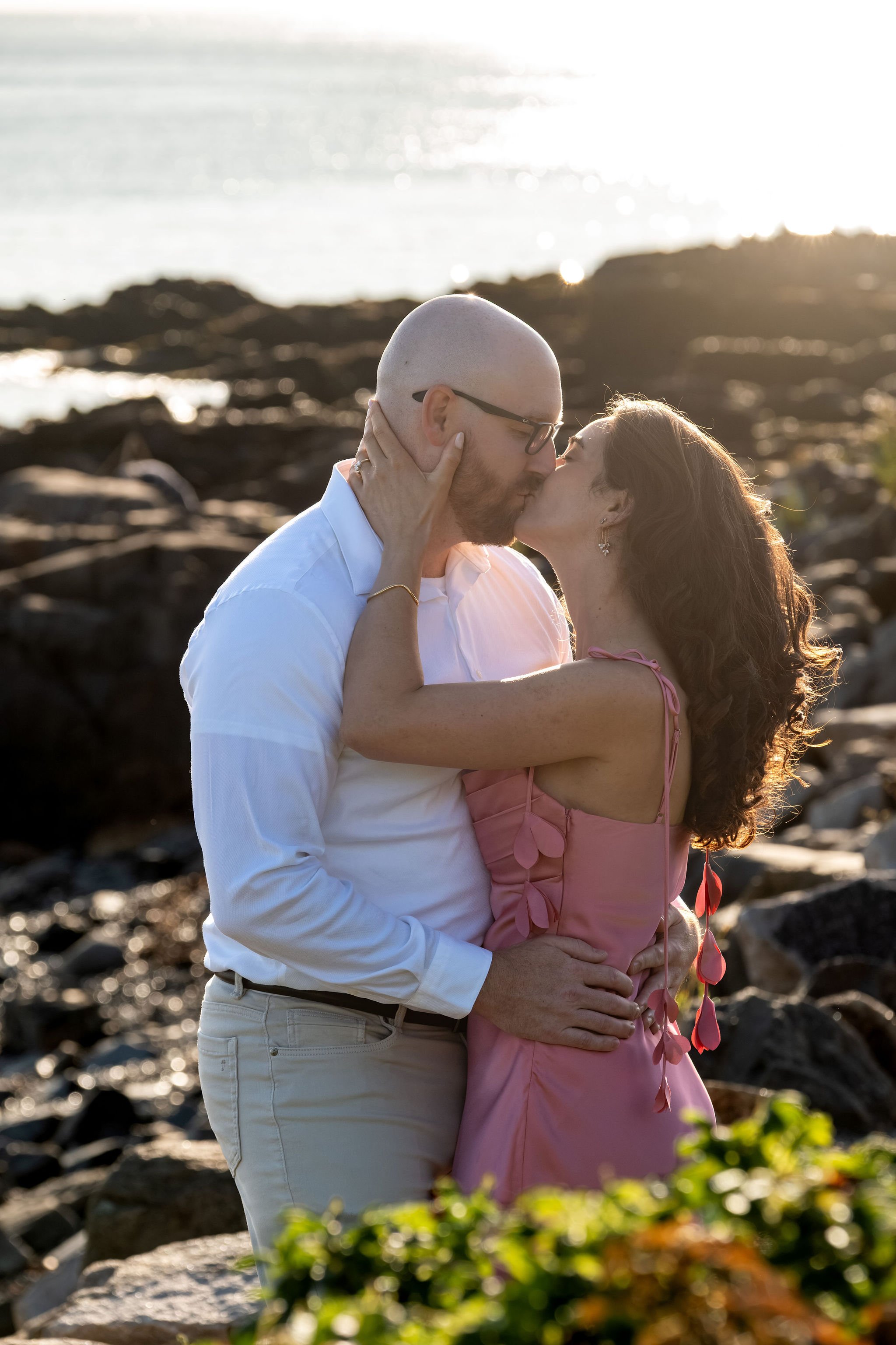 Zack and Helena kissing with the golden sunlight lighting up her curls on Marginal Way in Ogunquit Maine sunrise proposal and engagement photography VMG Productions