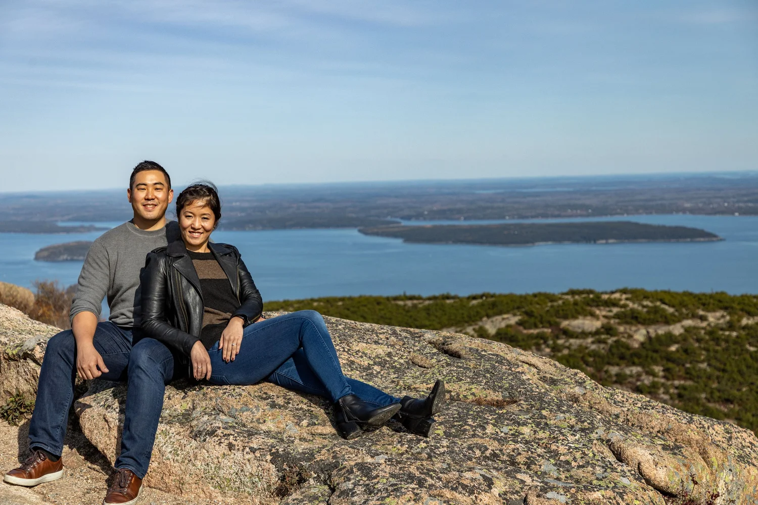 Agnes and Peter sitting on Cadillac Mountain summit smiling Blue Hill Bay Acadia National Park engagement VMG Productions