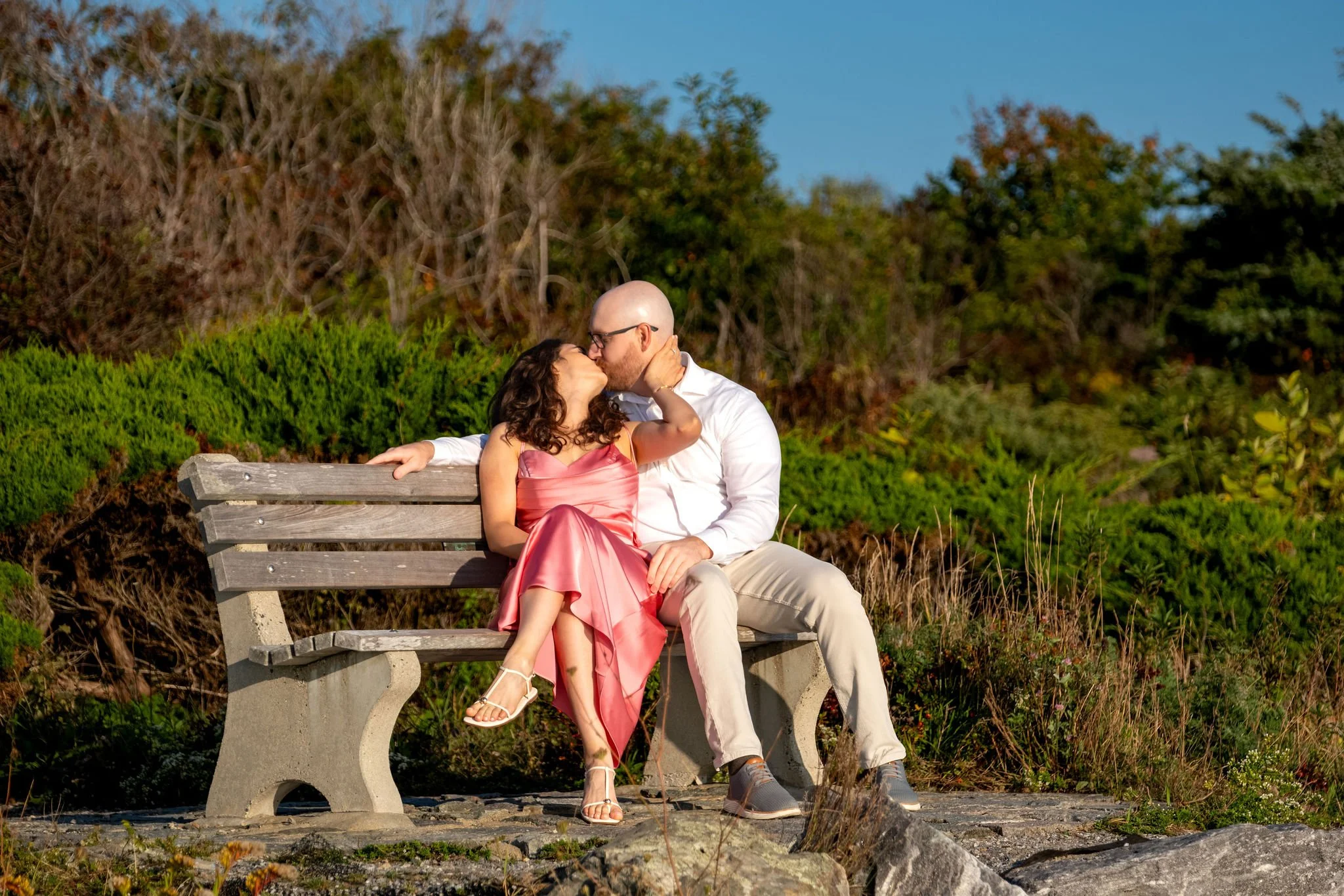 Zack and Helena sharing a kiss on the bench with the coastal brush behind them on Marginal Way in Ogunquit Maine sunrise proposal and engagement photography VMG Productions