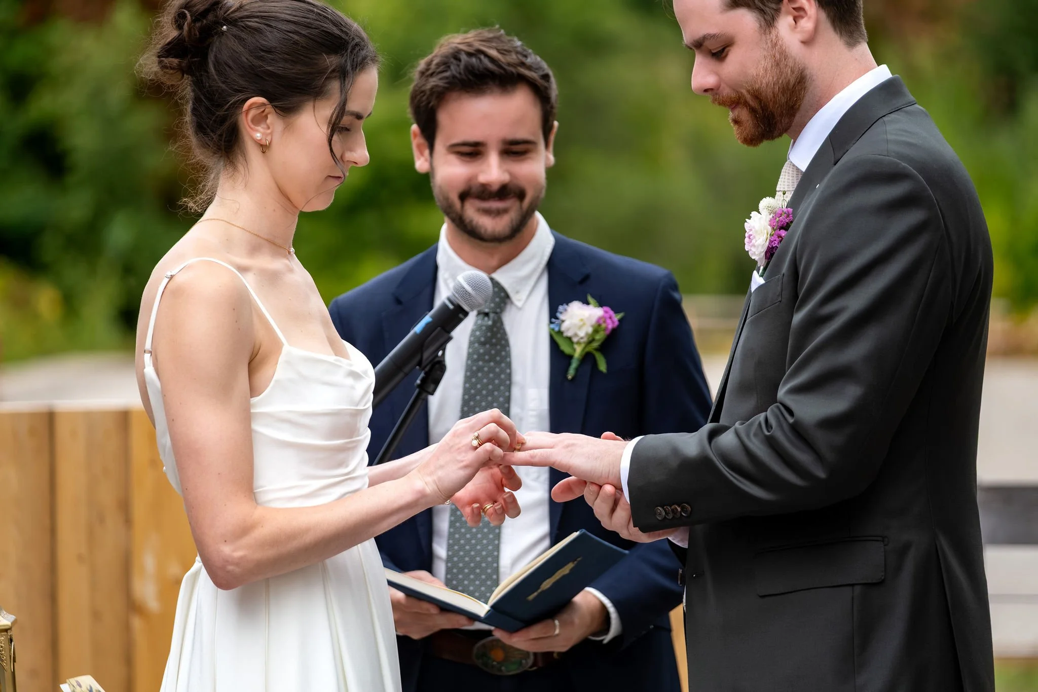 Victoria sliding the ring onto Jack's finger as the officiant smiles at The Cellars at Allagash Brewing Company in Portland Maine fall wedding photography VMG Productions