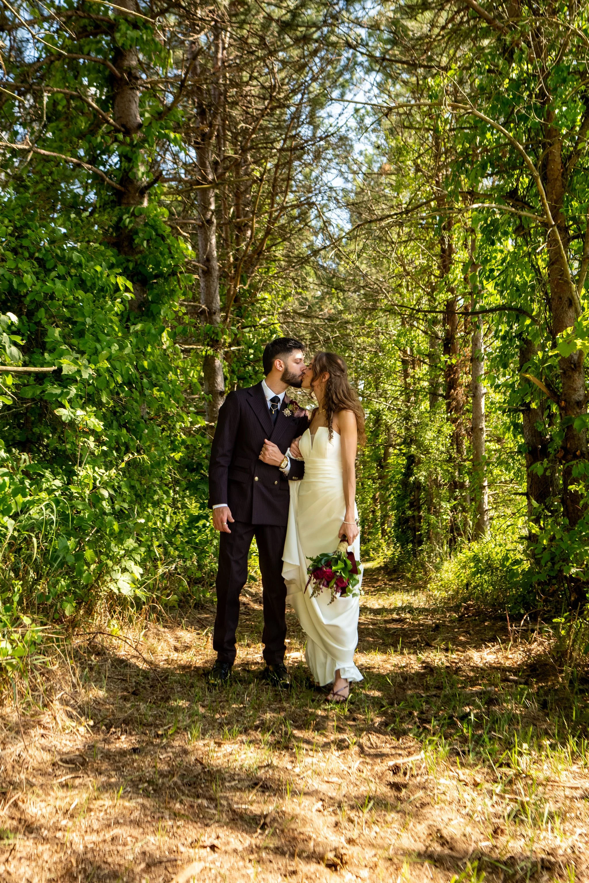 Cassidy and Chase Begin kissing on the pine trail with sunlight through the trees at Maple Hill Farm Inn and Conference Center in Hallowell Maine summer wedding photography VMG Productions