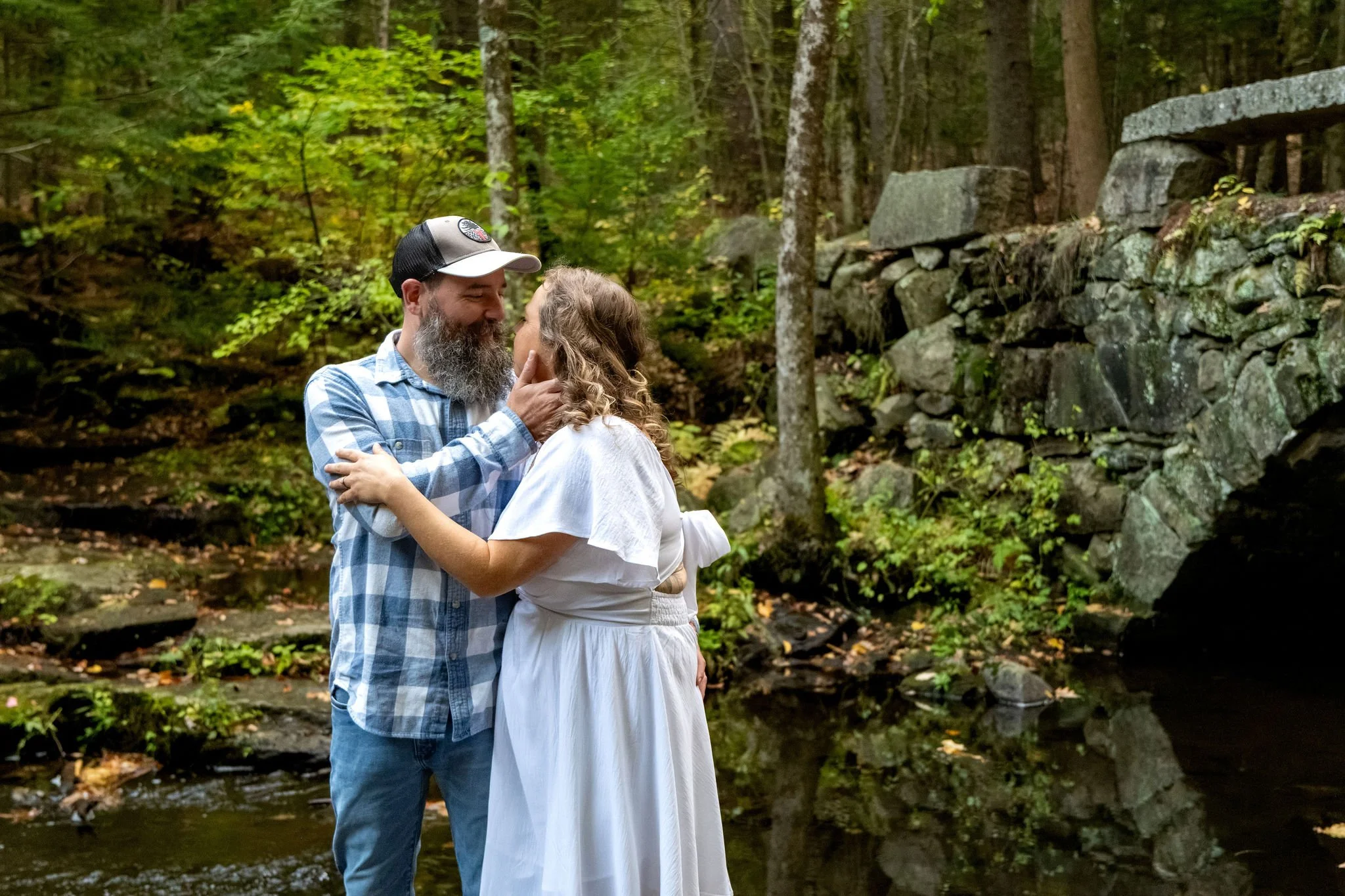 Alison and Dave standing close beside the stone wall with the brook and trees reflected in the still water at Vaughan Woods in Hallowell Maine fall engagement photography VMG Productions