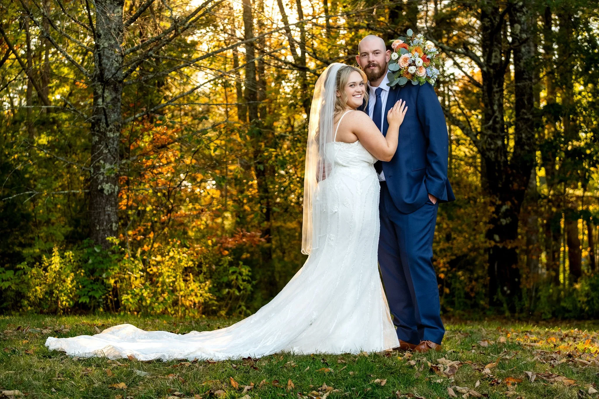 Ellie looking back at the camera in Sam's arms with her long train trailing across the grass and fall leaves at Boothbay Playhouse in Boothbay Maine fall wedding photography VMG Productions