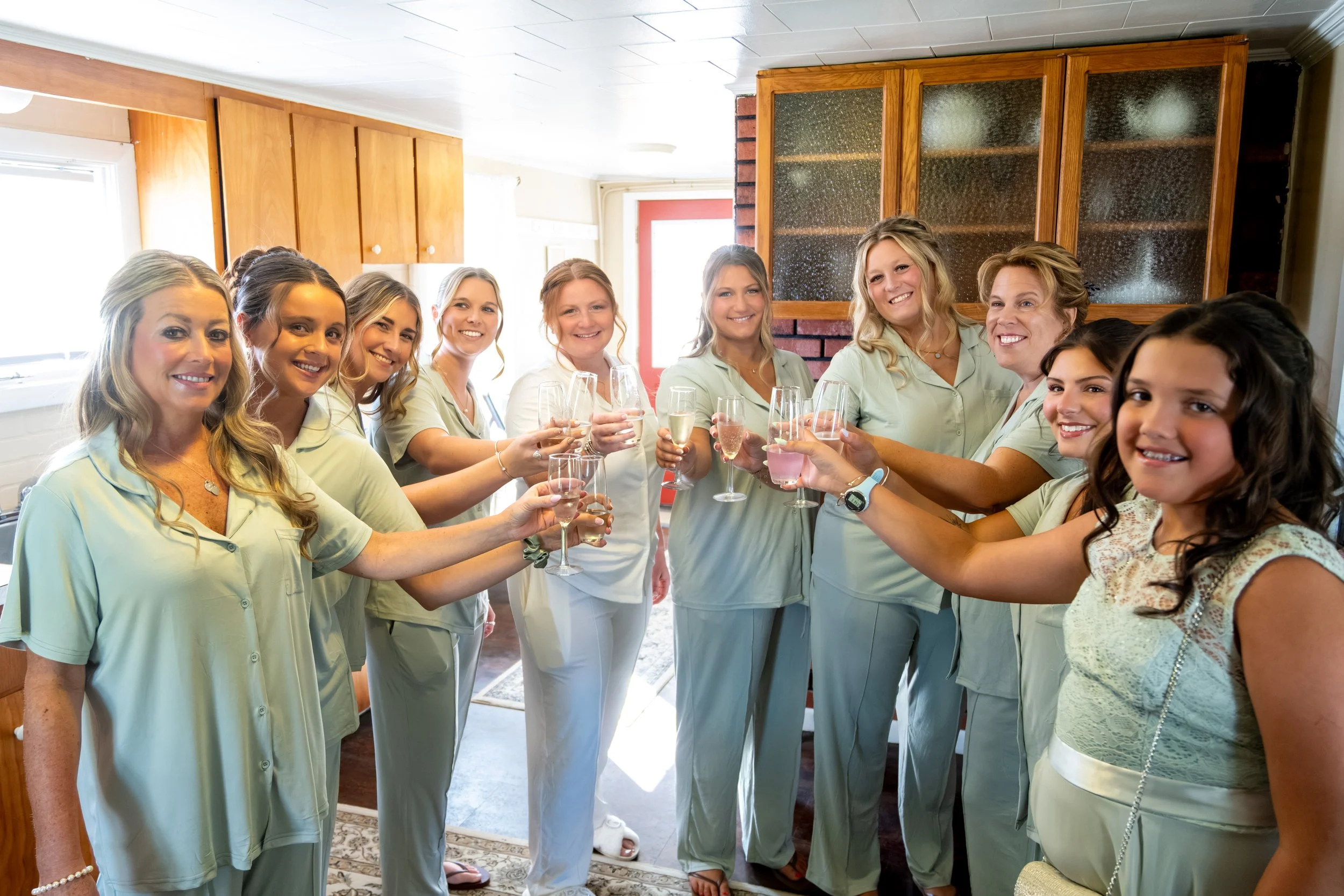 Erika's bridesmaids in sage pajamas raising their glasses together before the wedding at the Maine Wedding Barn and Event Center in Minot Maine fall wedding photography VMG Productions