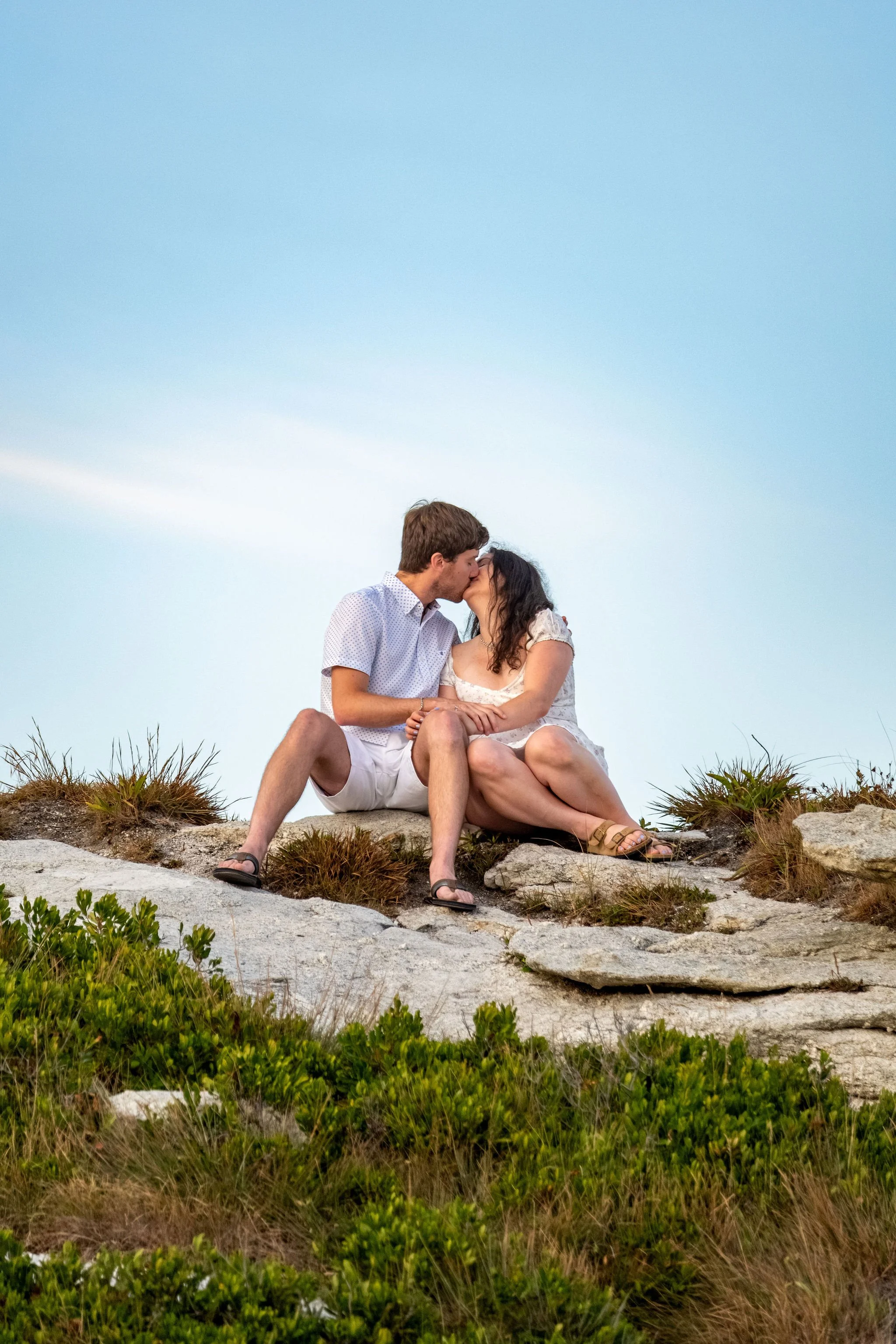 Kirstie and Lucas nose to nose seated on the rocks with blue sky behind them at Reid State Park in Georgetown Maine engagement photography VMG Productions