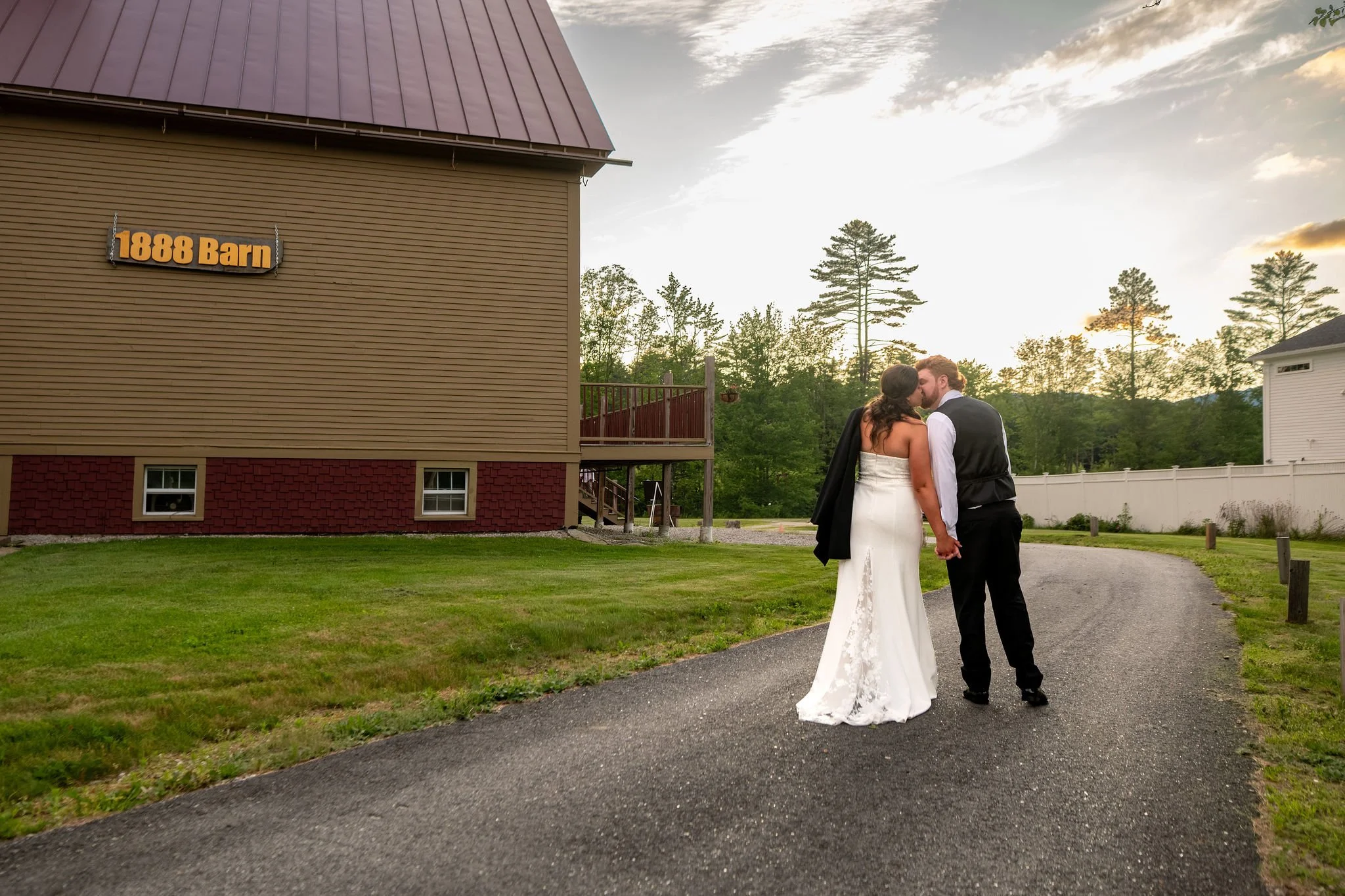 Alyssa and Colby kissing on the drive beneath the 1888 Barn sign as the sun sets behind the trees in Bethel Maine summer wedding photography VMG Productions