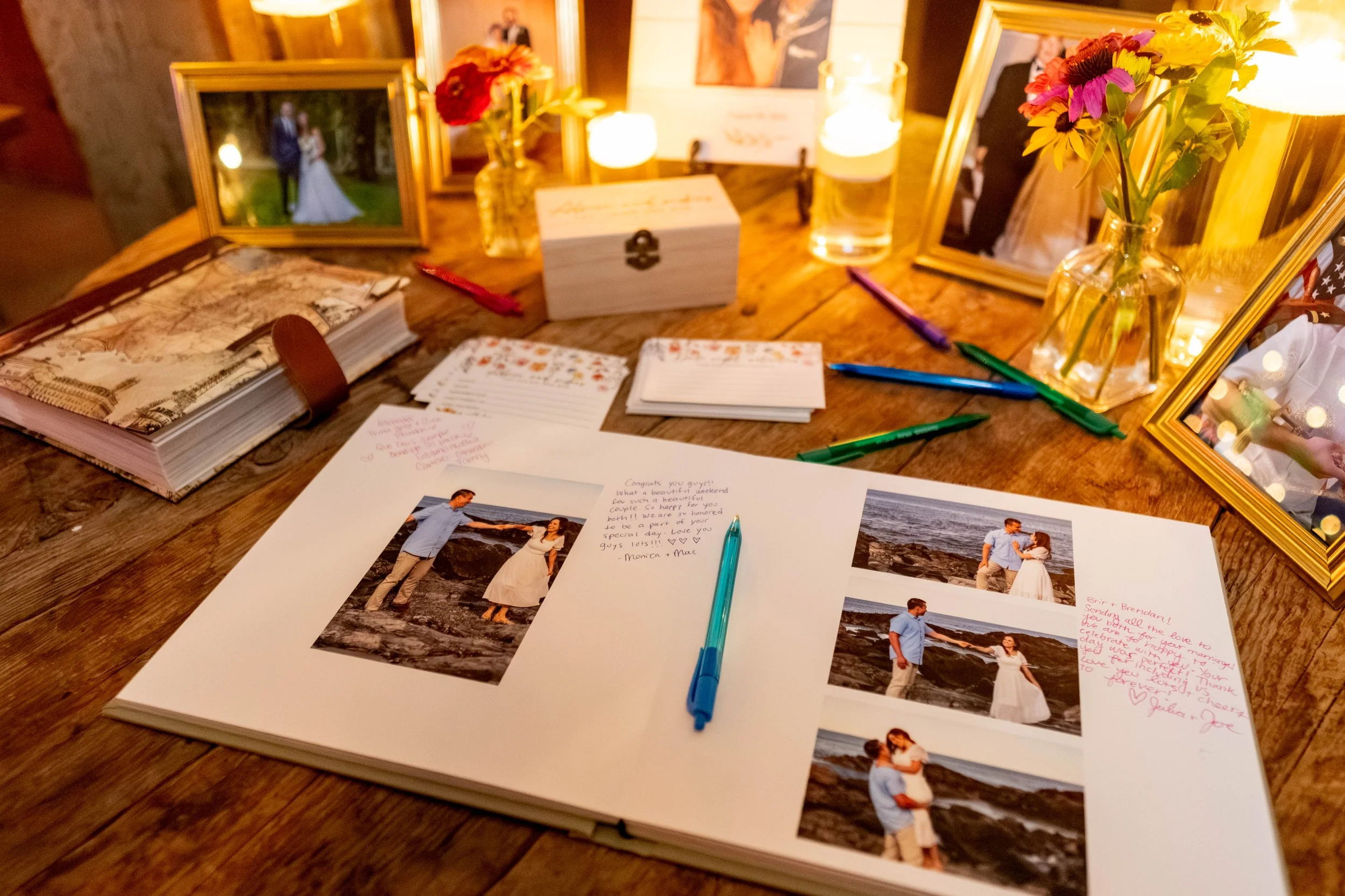 Printed wedding guest book album open showing multiple engagement session photos of couple on Maine rocky coast with handwritten guest messages filling white space around images at candlelit barn reception table