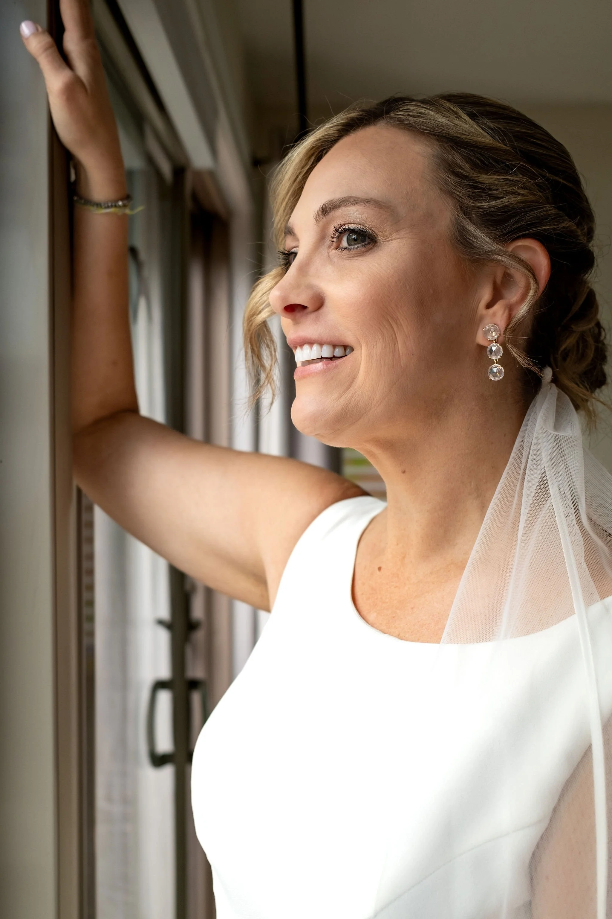 Alli Baudoin smiling in profile at the window in her veil while getting ready at a private residence in Mount Desert Maine spring wedding photography VMG Productions