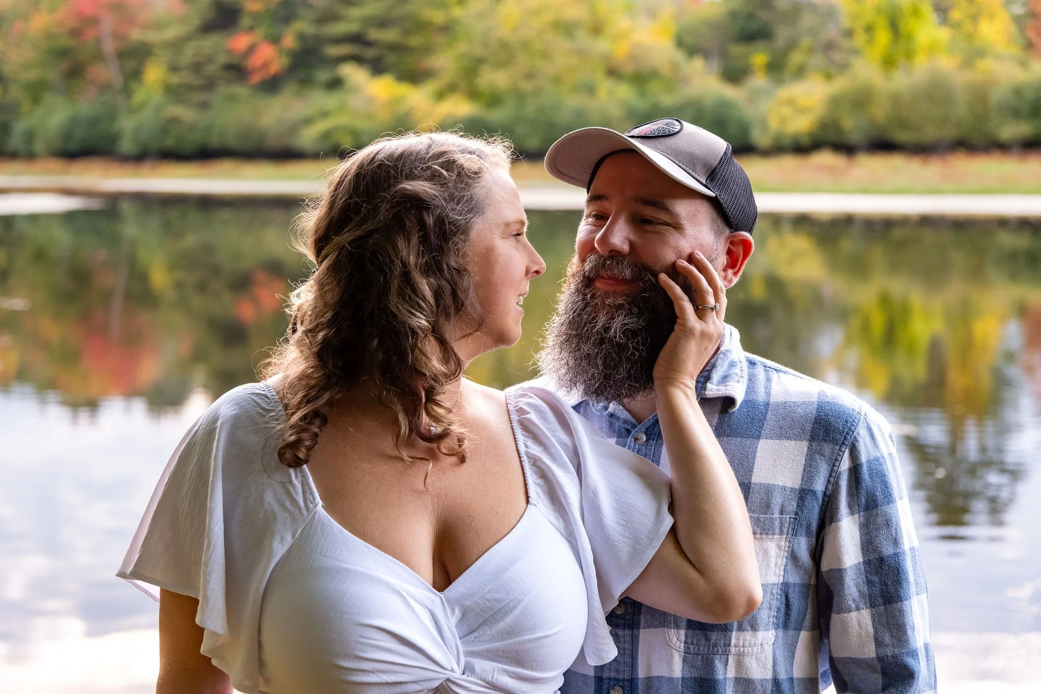 Alison holding Dave's cheek as they smile at each other with the fall trees reflected in the pond behind them at Vaughan Woods in Hallowell Maine fall engagement photography VMG Productions