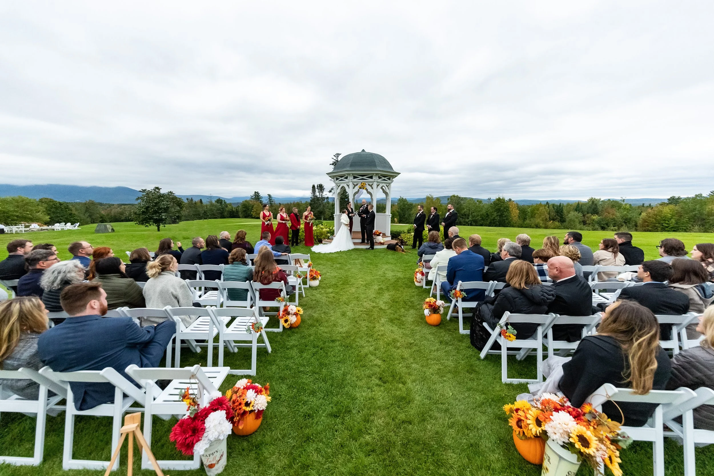 Wide outdoor ceremony shot at Mountain View Grand Resort Whitefield New Hampshire fall wedding 2022 showing guests seated on white folding chairs on green lawn facing white gazebo with couple and wedding party under cloudy mountain sky with fall foliage pumpkins and sunflower aisle arrangements