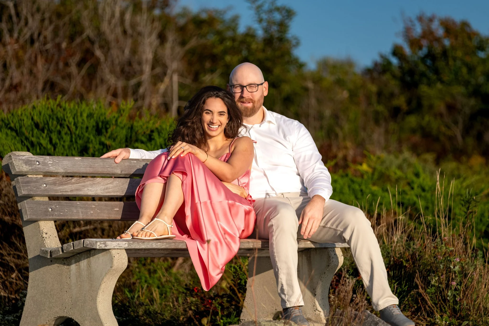 Helena smiling with her legs pulled up on the bench as Zack's arm rests along the back on Marginal Way in Ogunquit Maine sunrise proposal and engagement photography VMG Productions