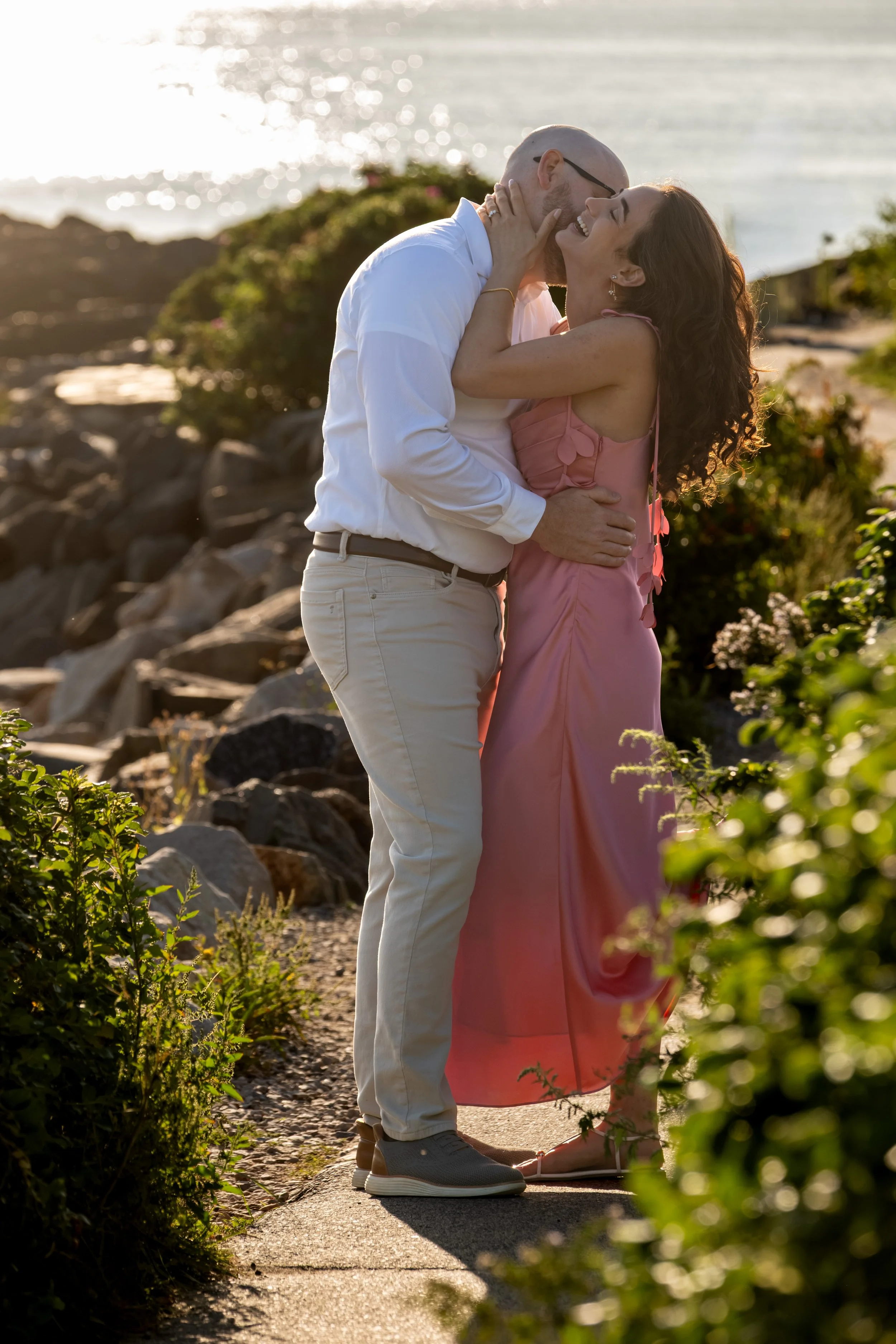 Zack and Helena laughing and embracing on the path with sunlight sparkling on the water behind them on Marginal Way in Ogunquit Maine sunrise proposal and engagement photography VMG Productions