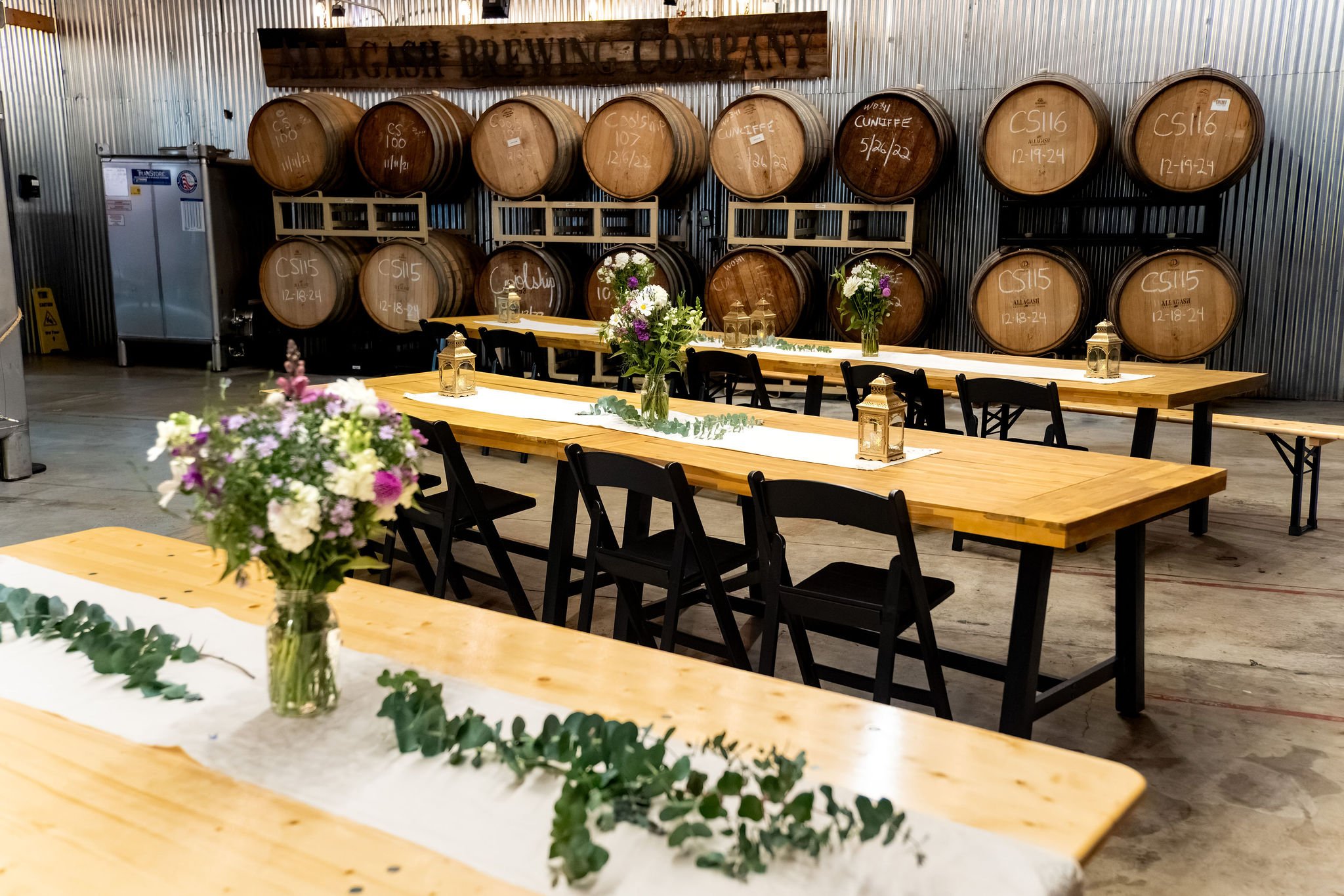 Wooden reception tables set with jars of wildflowers, lanterns, and eucalyptus runners in front of oak barrels at The Cellars at Allagash Brewing Company in Portland Maine fall wedding photography VMG Productions