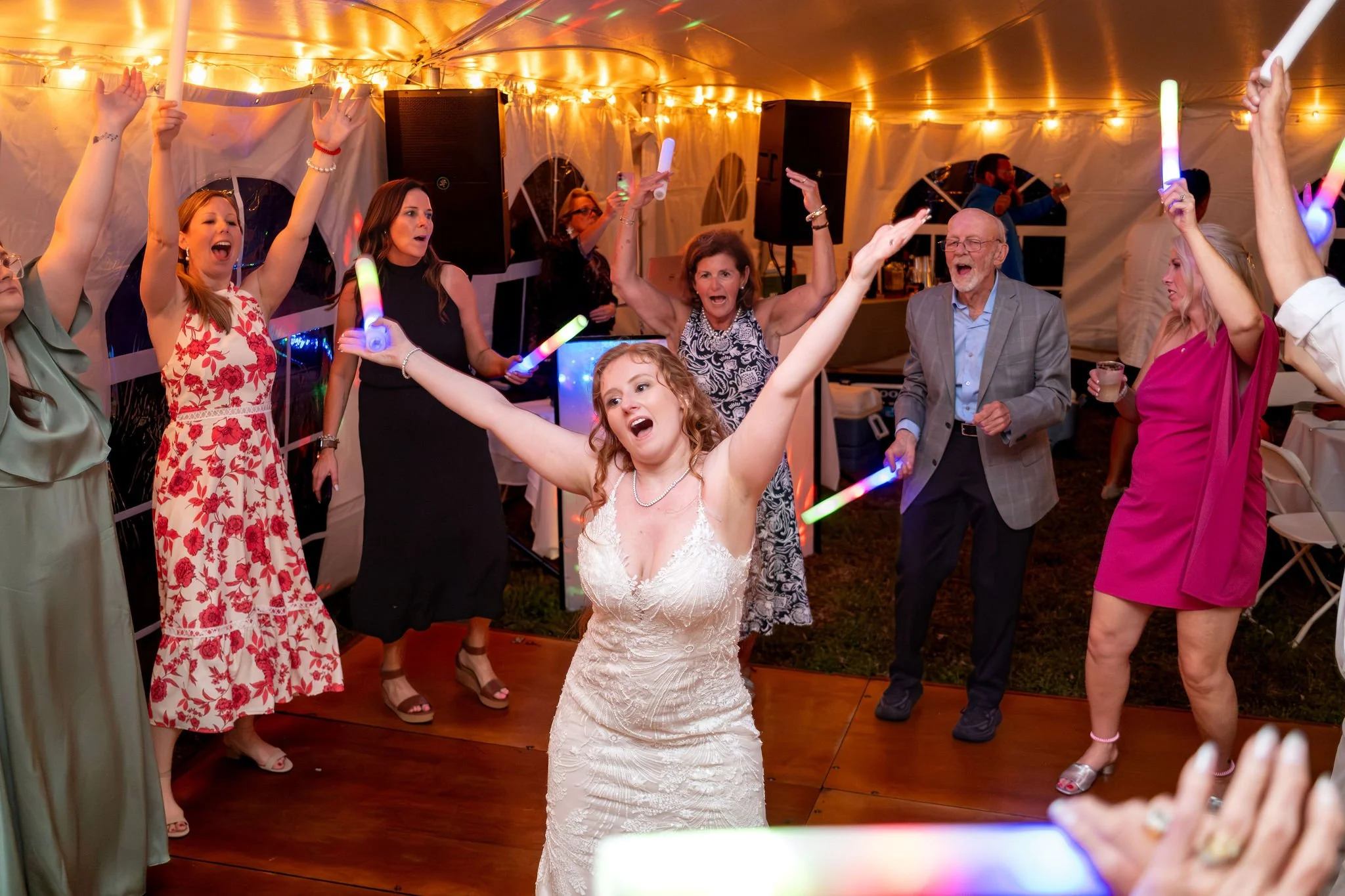 Kayla DeAngelis with both arms raised on the dance floor surrounded by guests with glow sticks inside the reception tent at their Sanbornton New Hampshire wedding photography VMG Productions