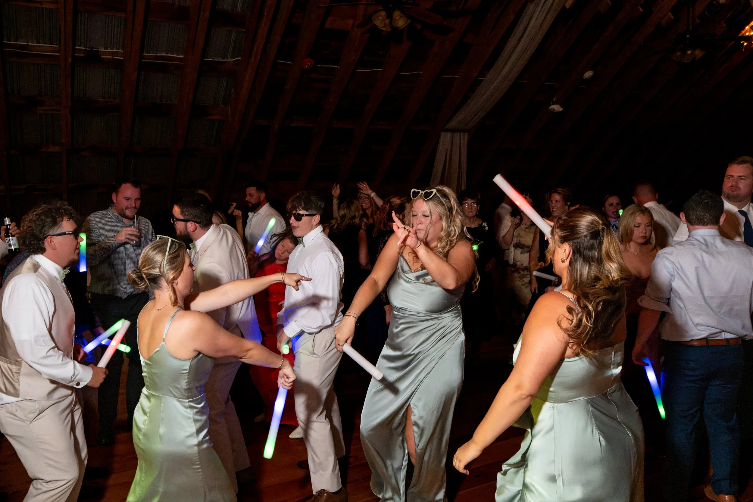 A packed dance floor of guests dancing with light-up wands under the barn rafters at the Maine Wedding Barn and Event Center in Minot Maine fall wedding photography VMG Productions