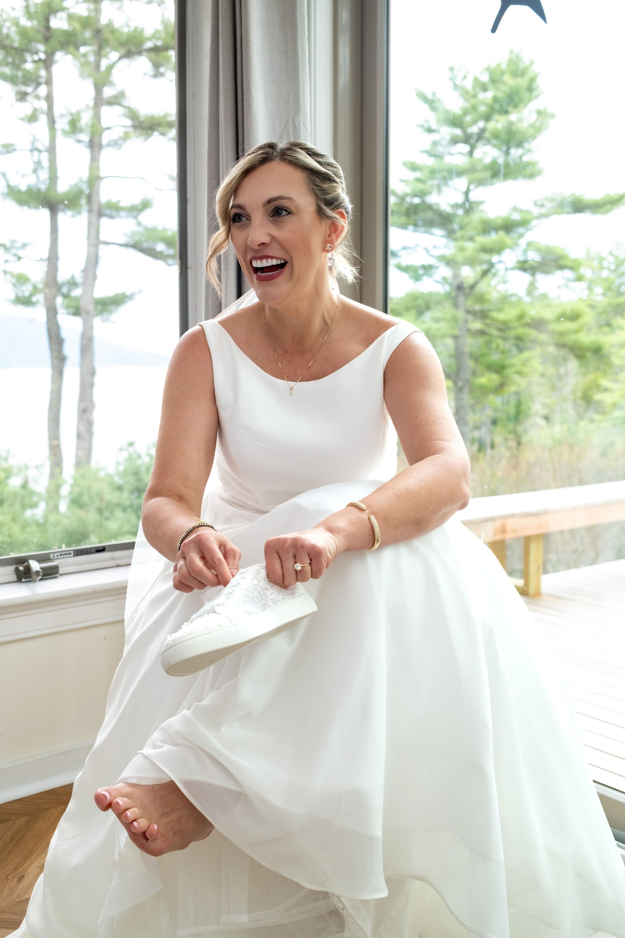 Alli Baudoin laughing while lacing her white lace sneakers by the window overlooking Somes Sound in Mount Desert Maine spring wedding photography VMG Productions