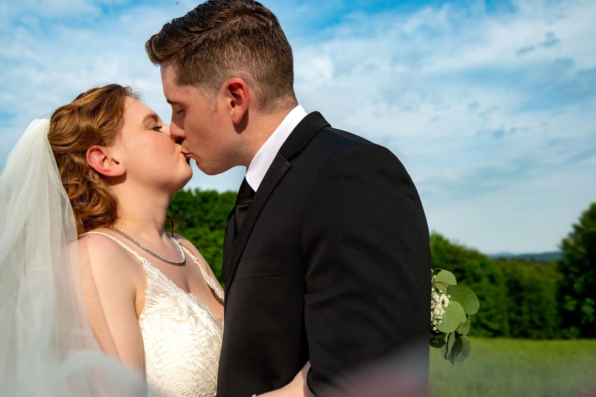 Kayla and Ethan DeAngelis kissing in the summer field with the New Hampshire hills behind them at their Sanbornton private residence wedding photography VMG Productions
