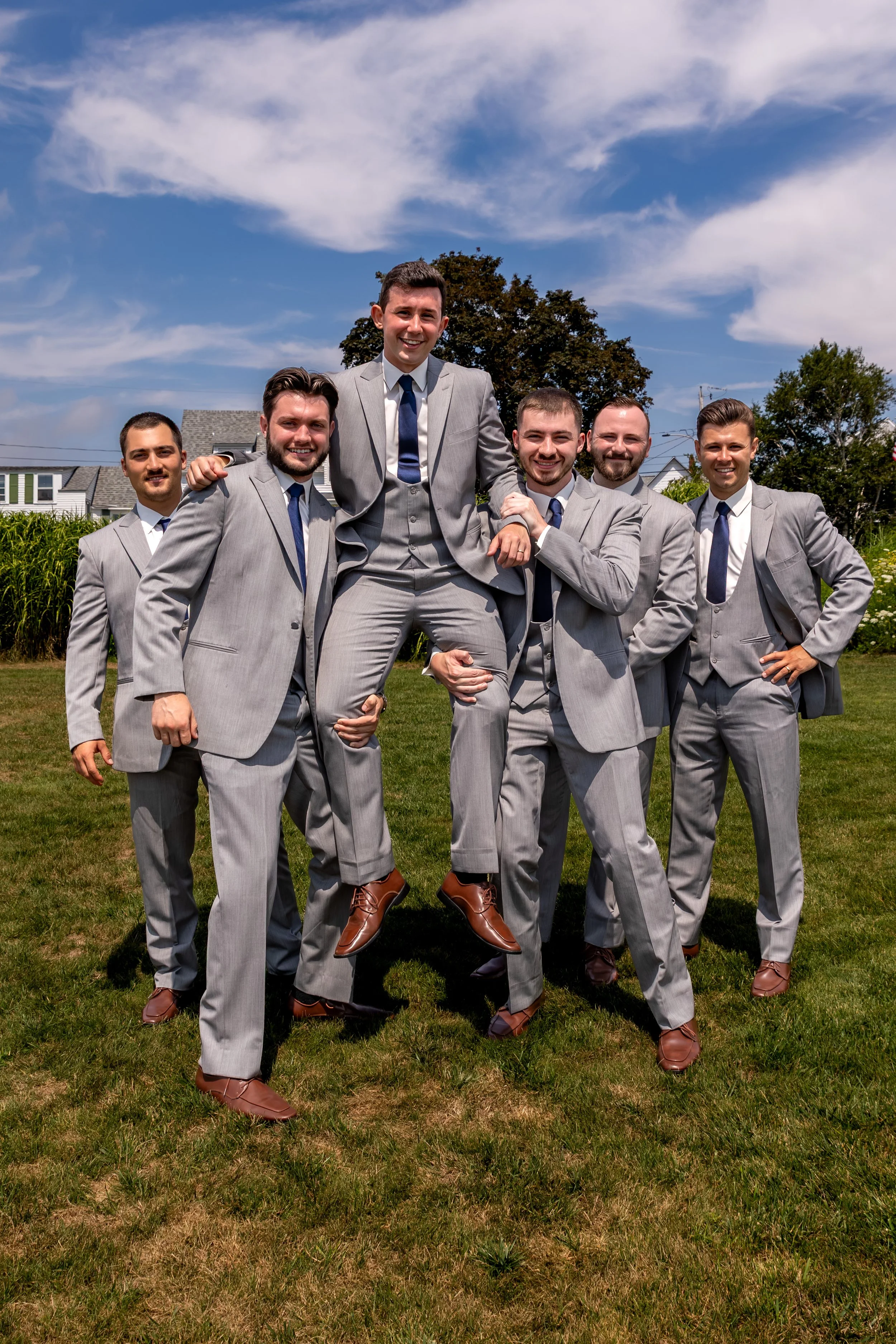 Anthony Pirolli's groomsmen in grey suits lifting him on the lawn at Union Bluff Hotel in York Maine wedding photography VMG Productions