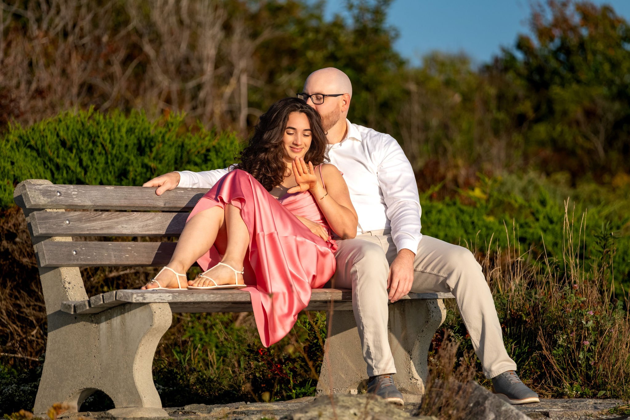 Helena tucked against Zack on the bench with her eyes closed and a soft smile on Marginal Way in Ogunquit Maine sunrise proposal and engagement photography VMG Productions