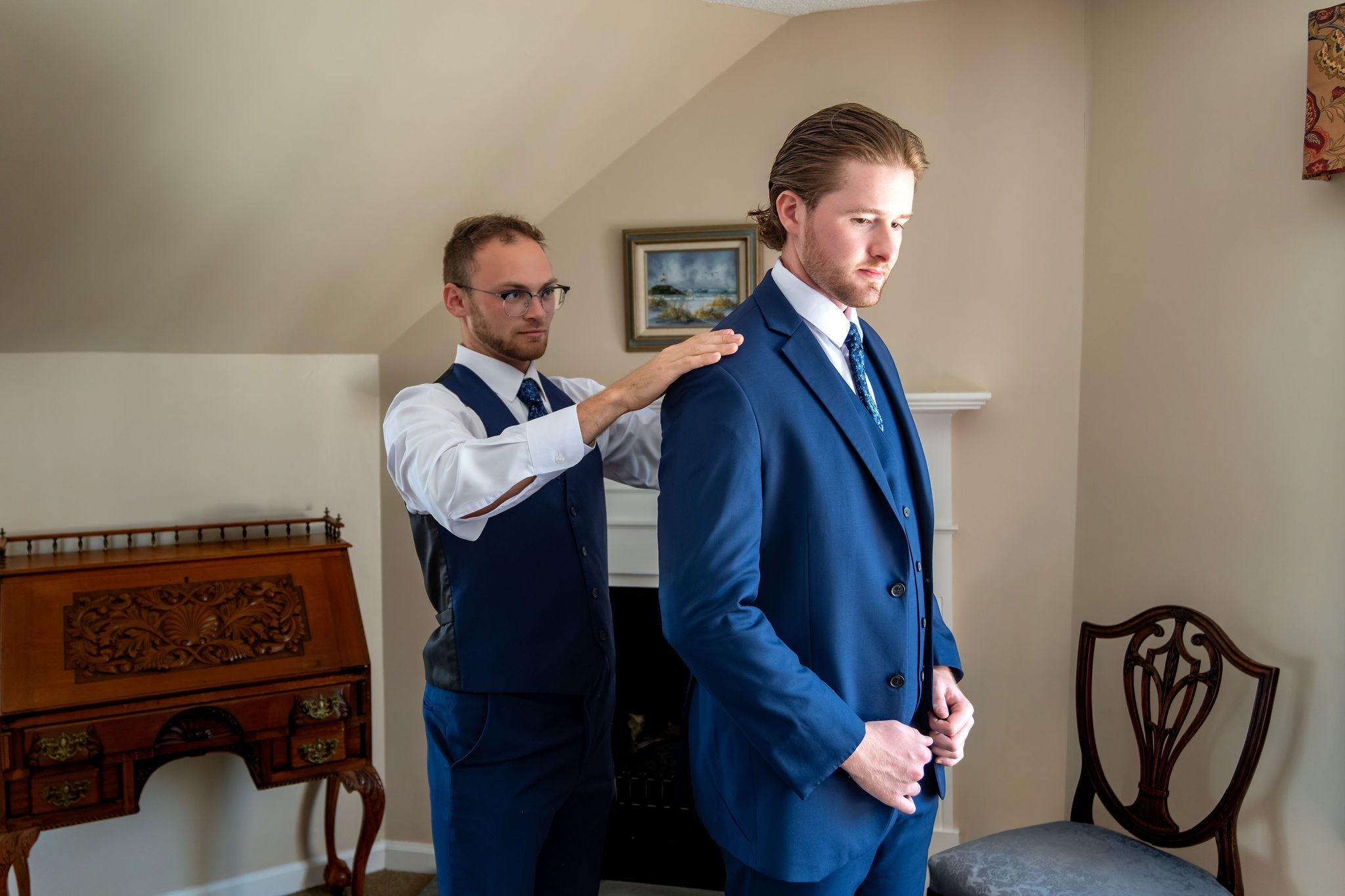 A groomsman helping Zachary into his navy suit jacket in front of the fireplace at The Lucerne Inn in Dedham Maine summer wedding photography VMG Productions